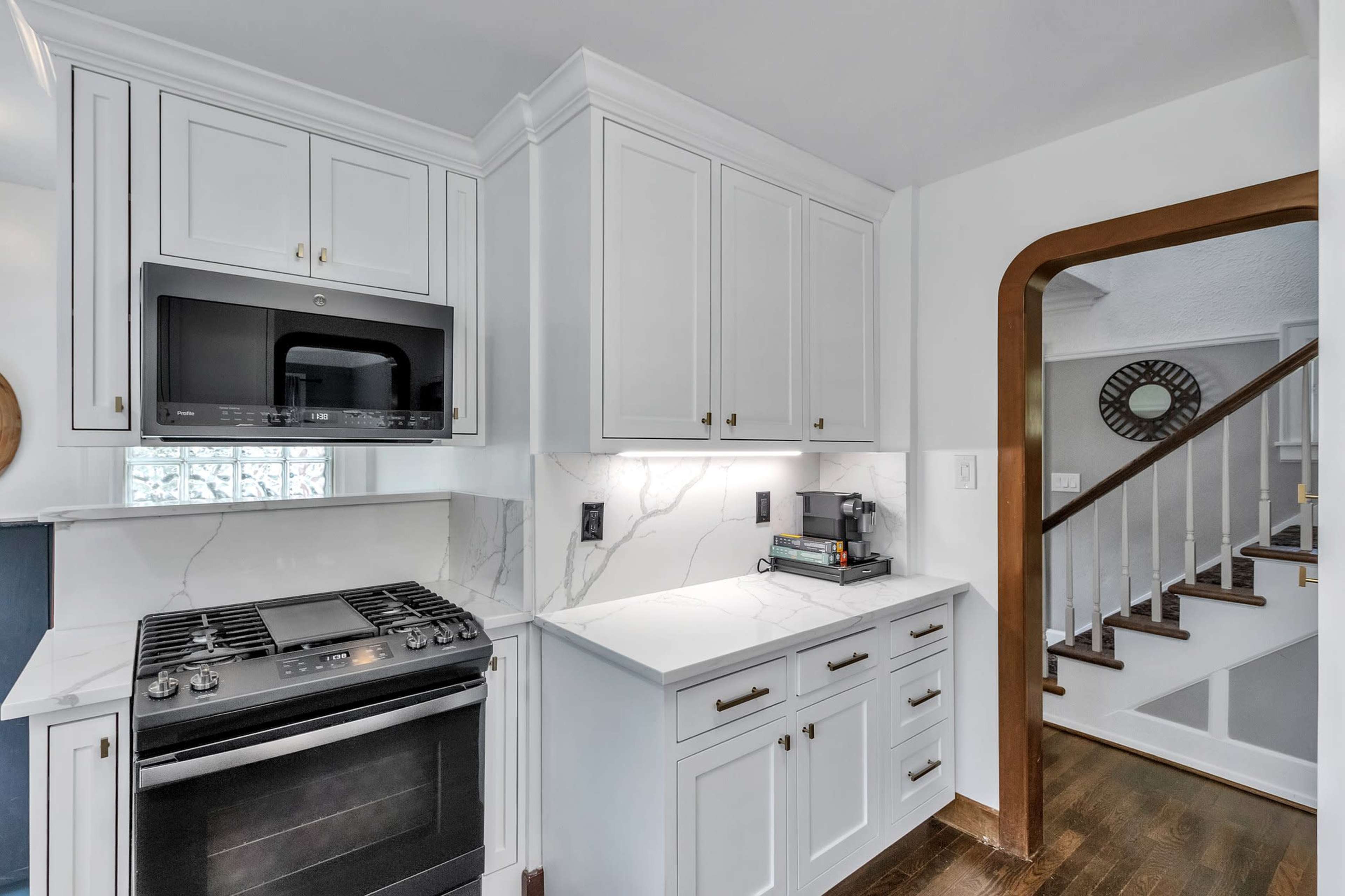 The image shows a modern kitchen with white cabinetry, a built-in microwave, and a gas stove, adjacent to an entryway leading to a staircase.