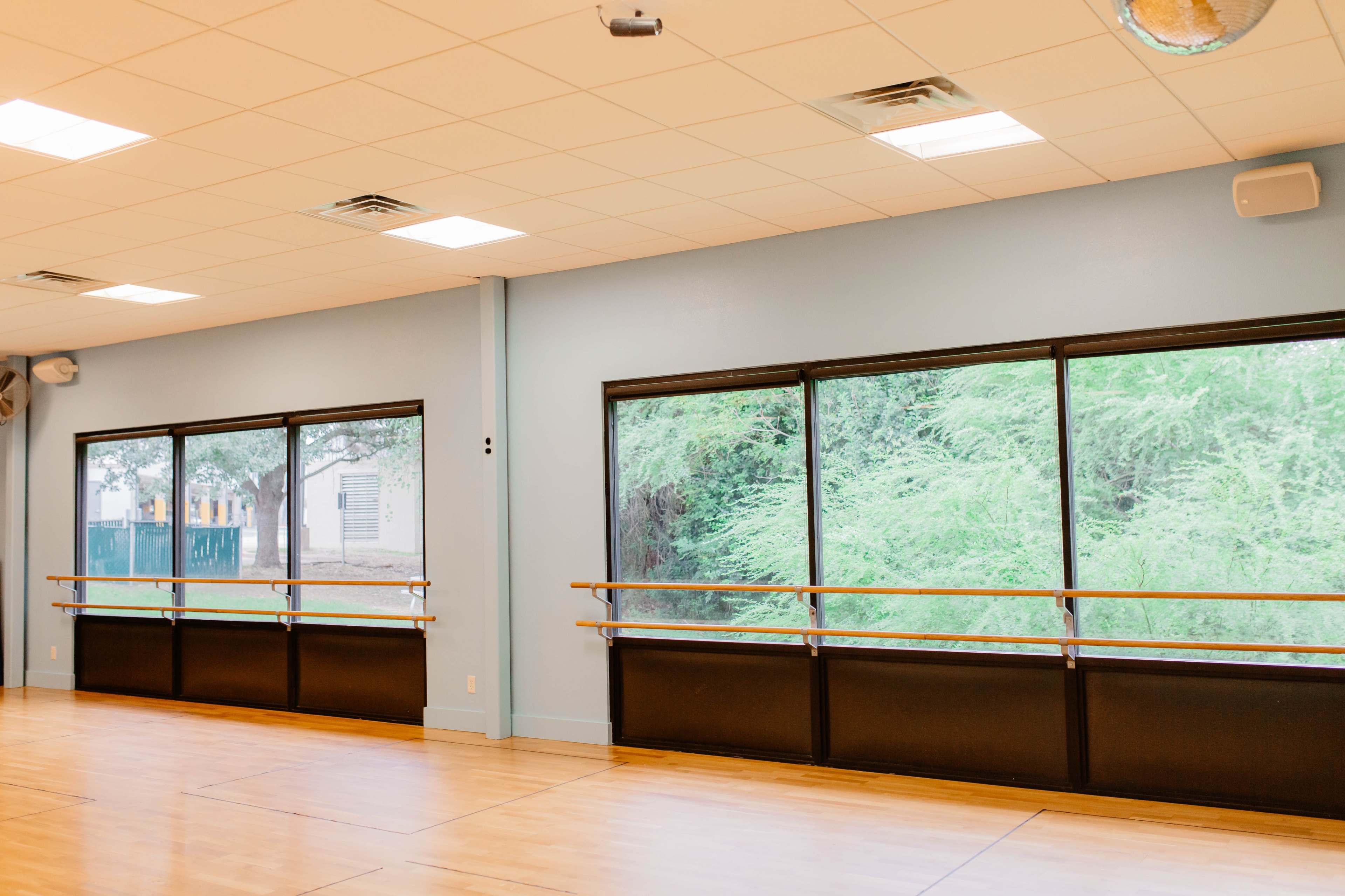 The image shows a dance studio with wooden flooring, large windows providing natural light, and ballet barres along the walls.