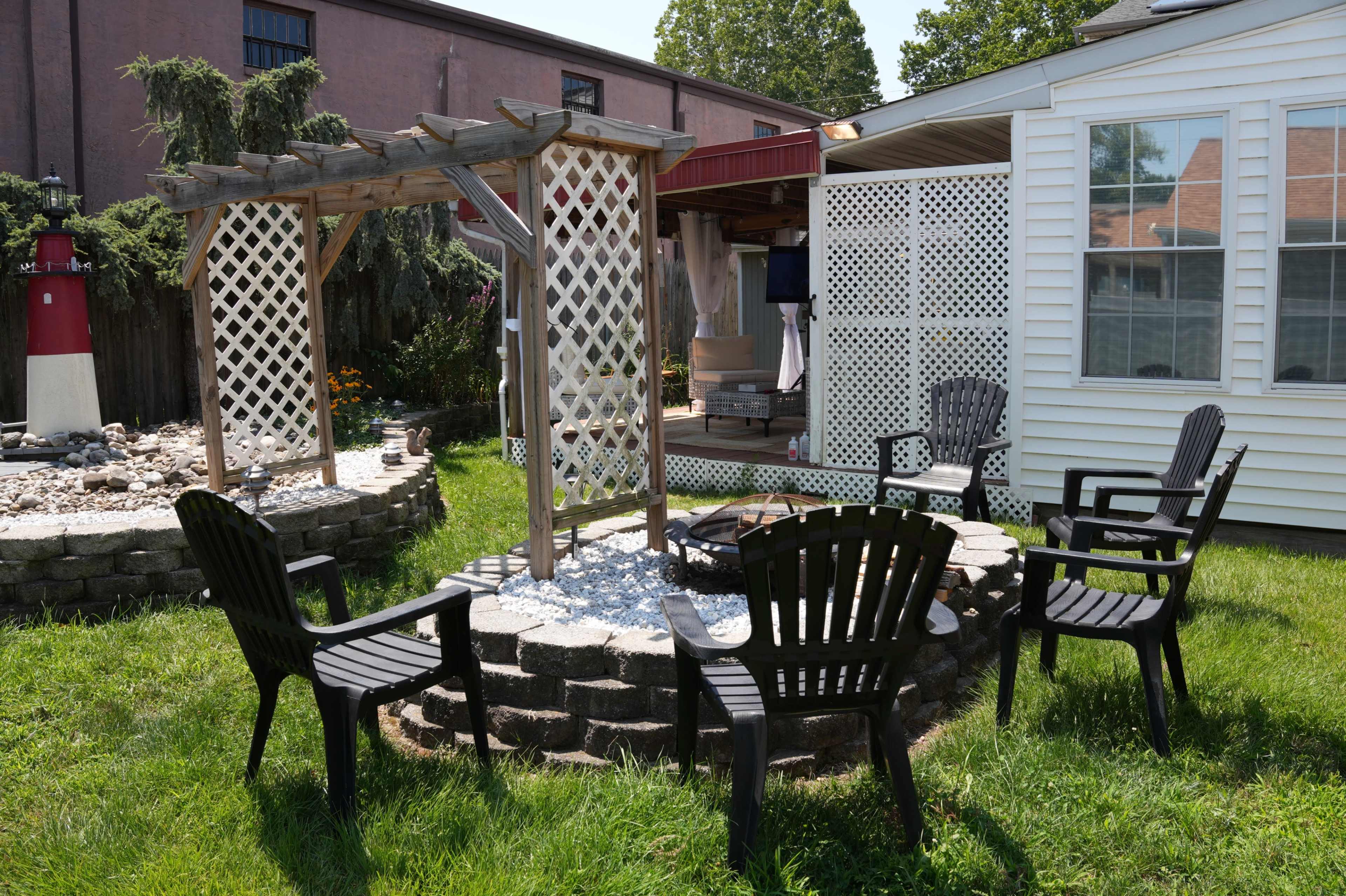 The image shows a landscaped backyard featuring a stone seating area with chairs around a circular fire pit and a wooden arbor beside a gravel bed.