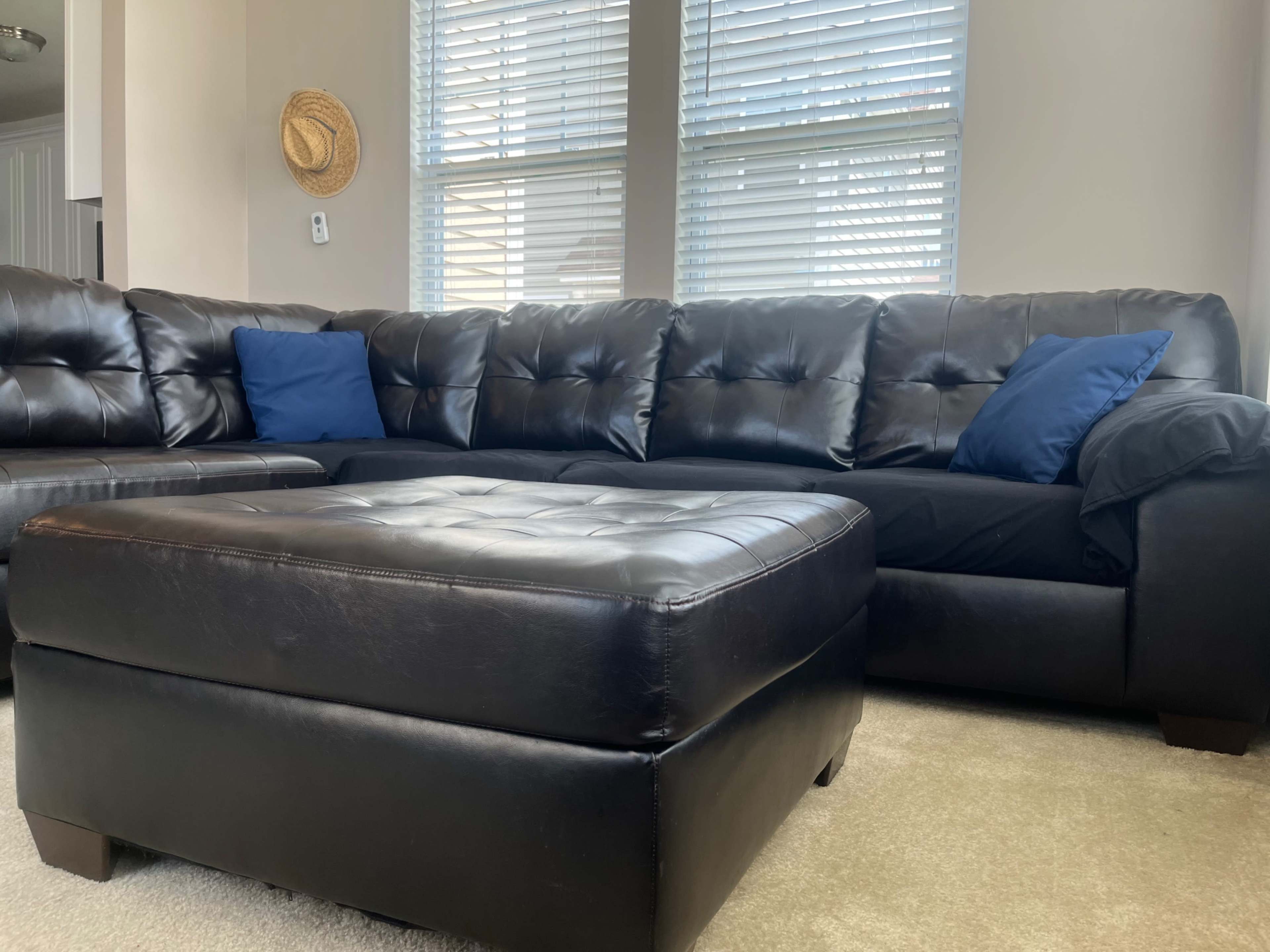 A black leather sectional sofa with blue throw pillows is positioned beside a large leather ottoman in a well-lit room.