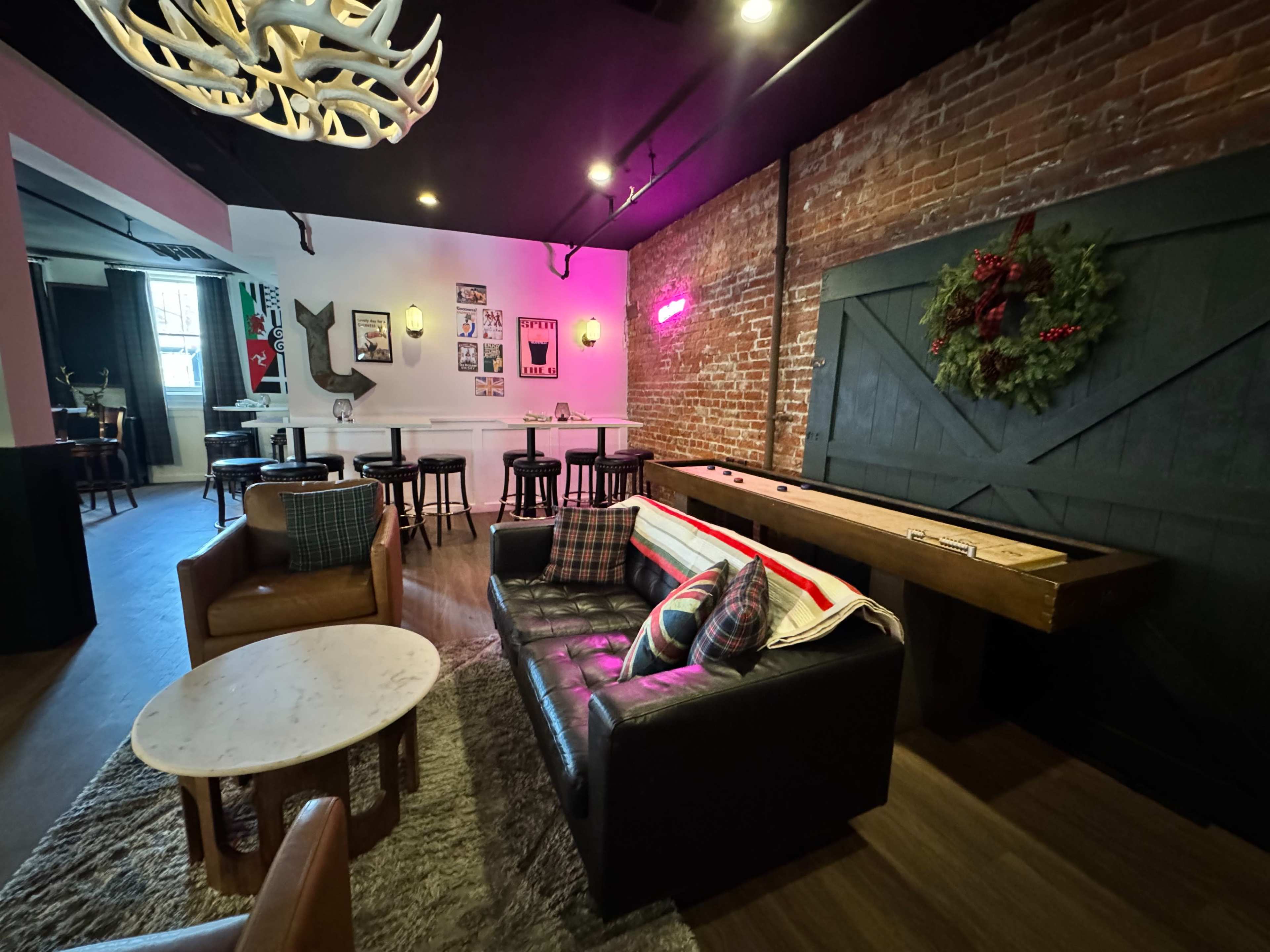 The image shows a cozy lounge area with a black leather sofa, a round marble table, and a shuffleboard table against a brick wall adorned with festive decorations.