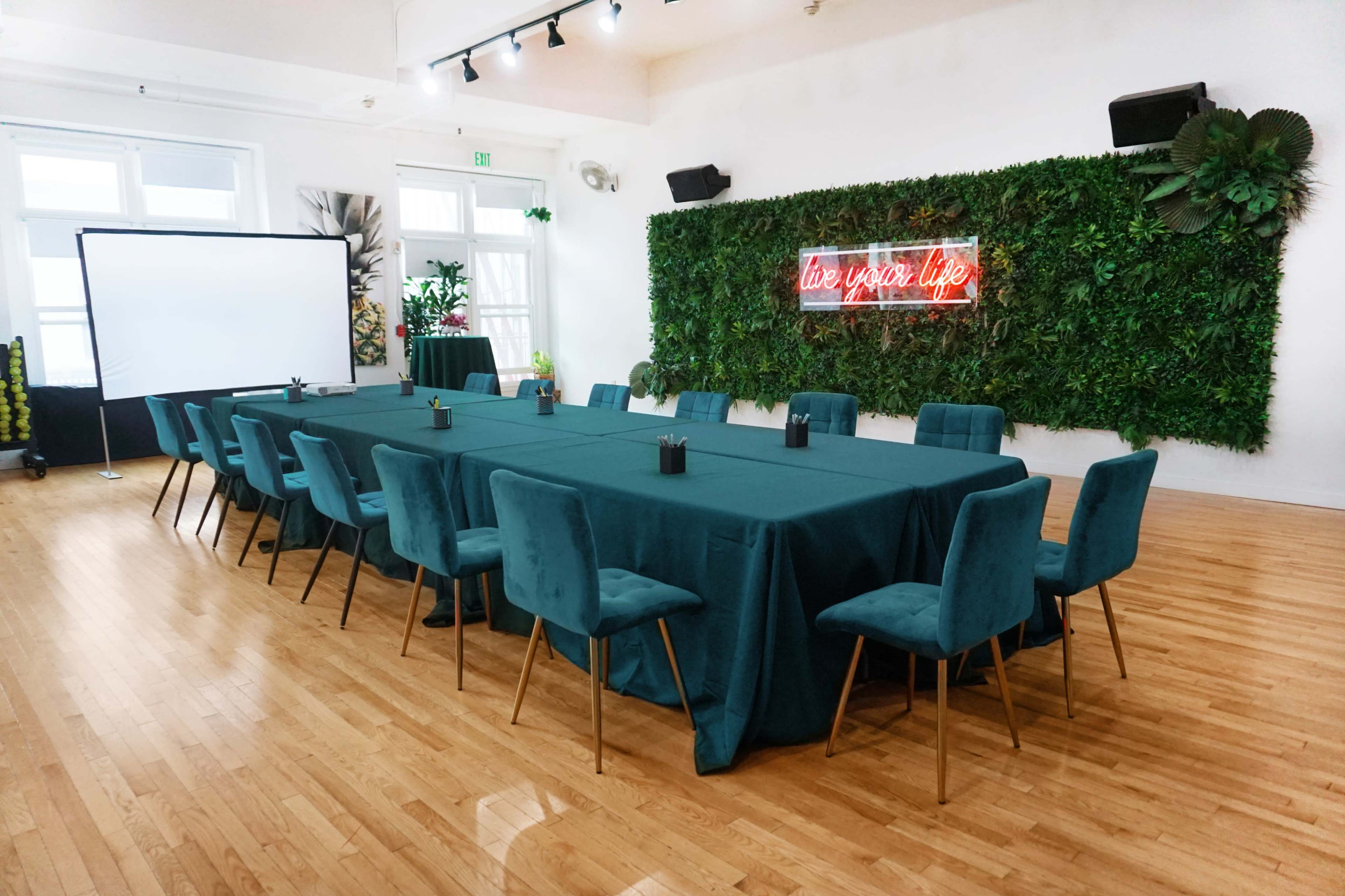 A large, rectangular conference table with teal tablecloths and matching chairs is set up in a brightly lit room featuring a neon sign and a vertical garden backdrop.