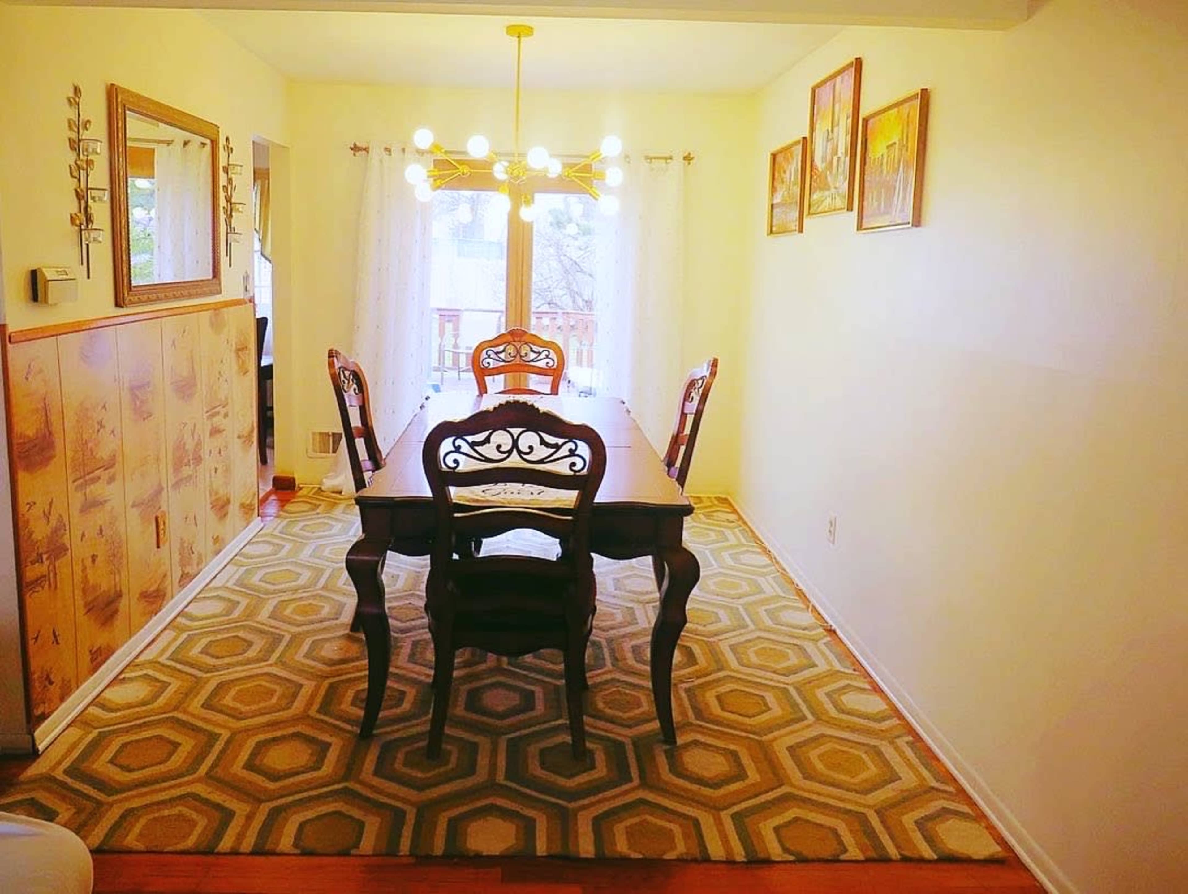 The image shows a dining room with a wooden table and four chairs, a patterned rug, and large windows letting in natural light.