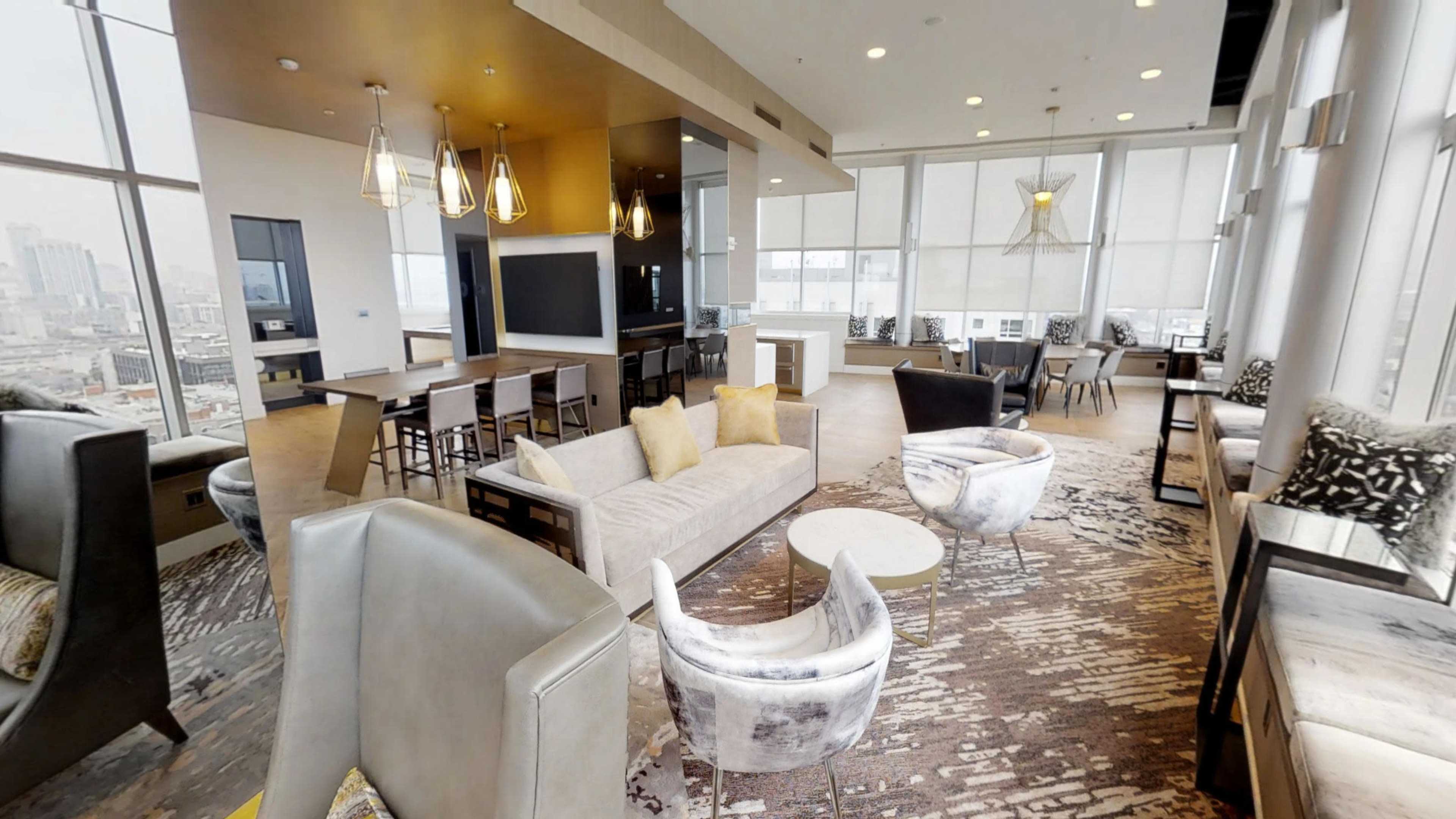 A modern lounge area features comfortable seating, large windows with city views, and a dining table.