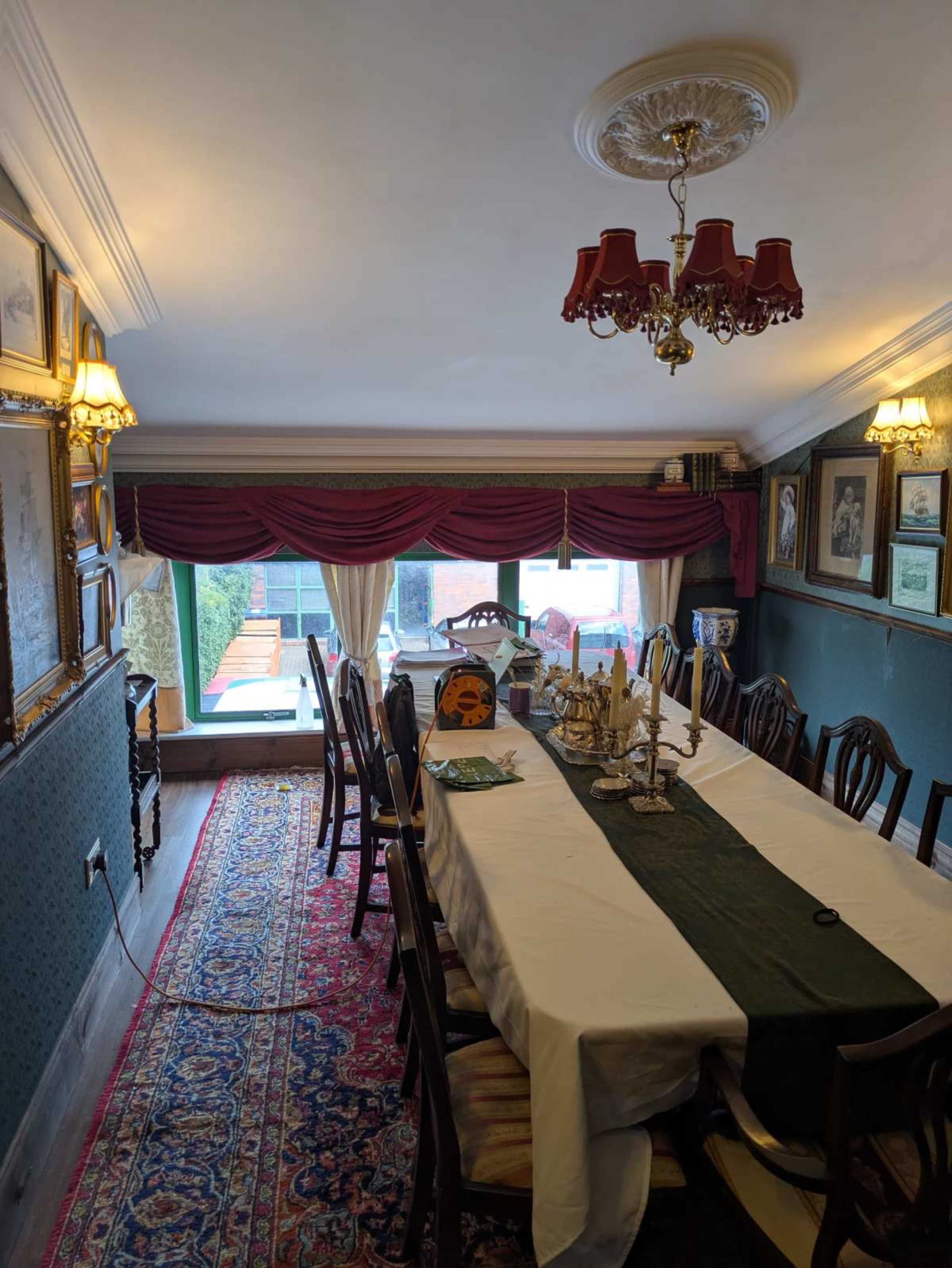 A long dining table is set with a green table runner in a well-decorated dining room featuring chandeliers and framed artworks on the walls.