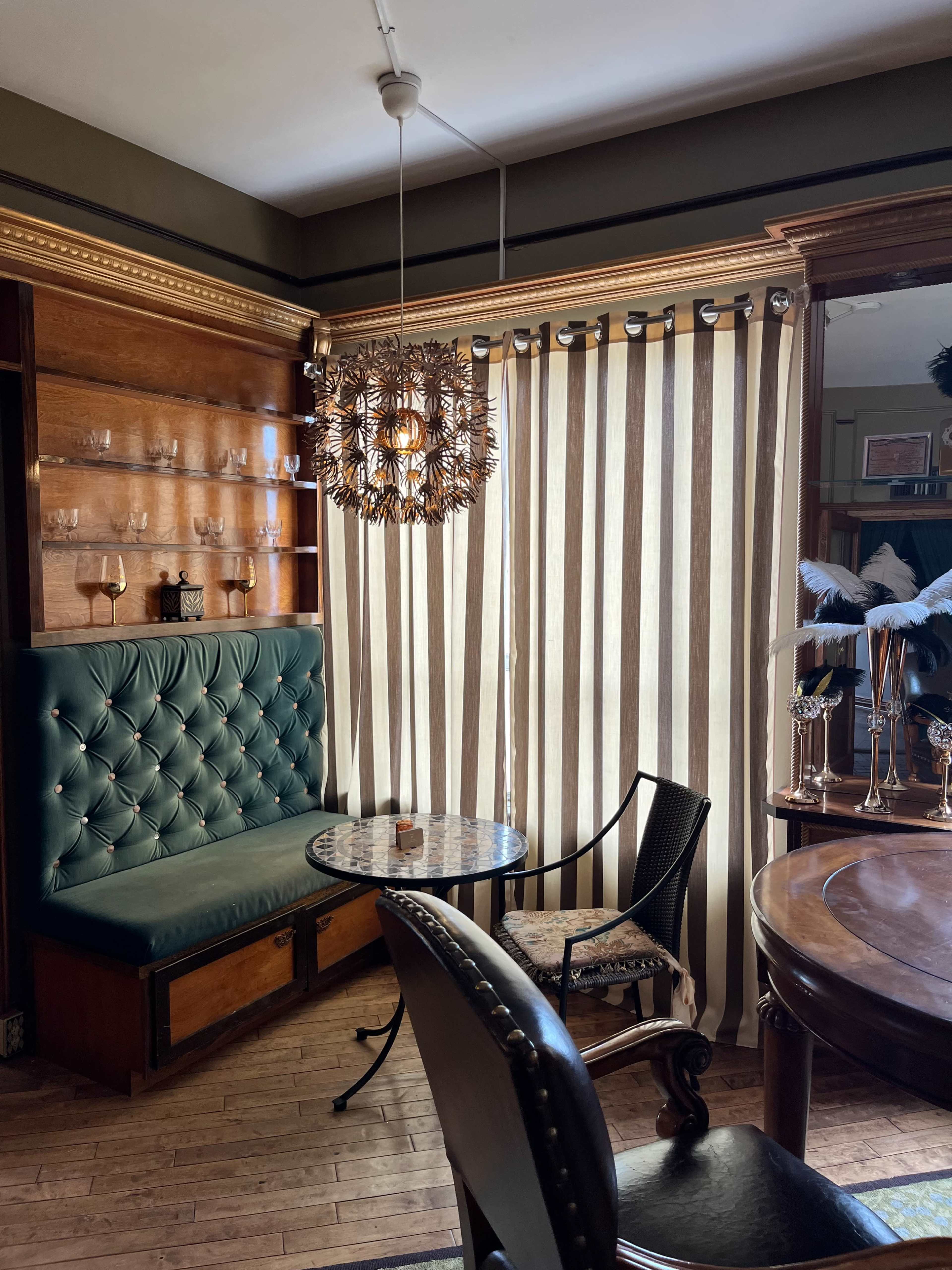 A cozy seating area with a green upholstered bench, a round table, and striped curtains beside a wooden bookshelf.
