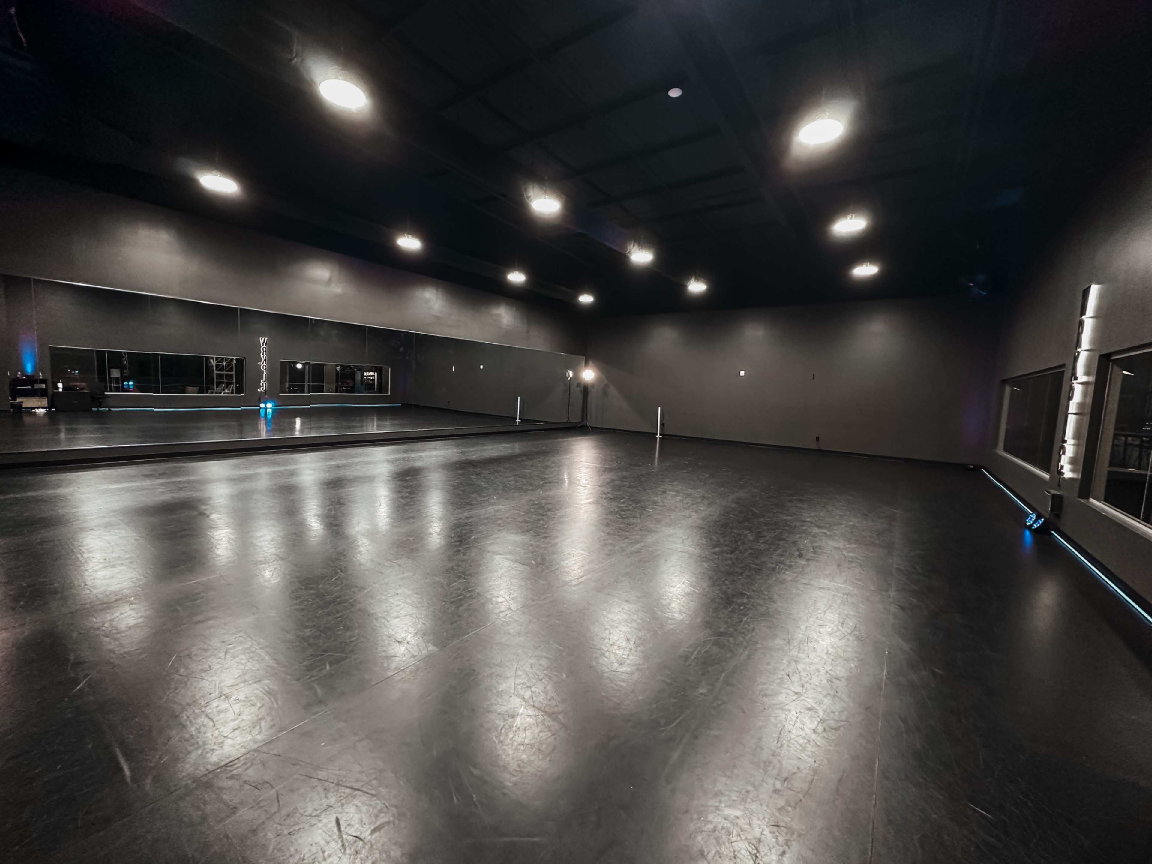 The image shows a spacious dance studio with a polished dark floor, mirrors along one wall, and dim lighting.