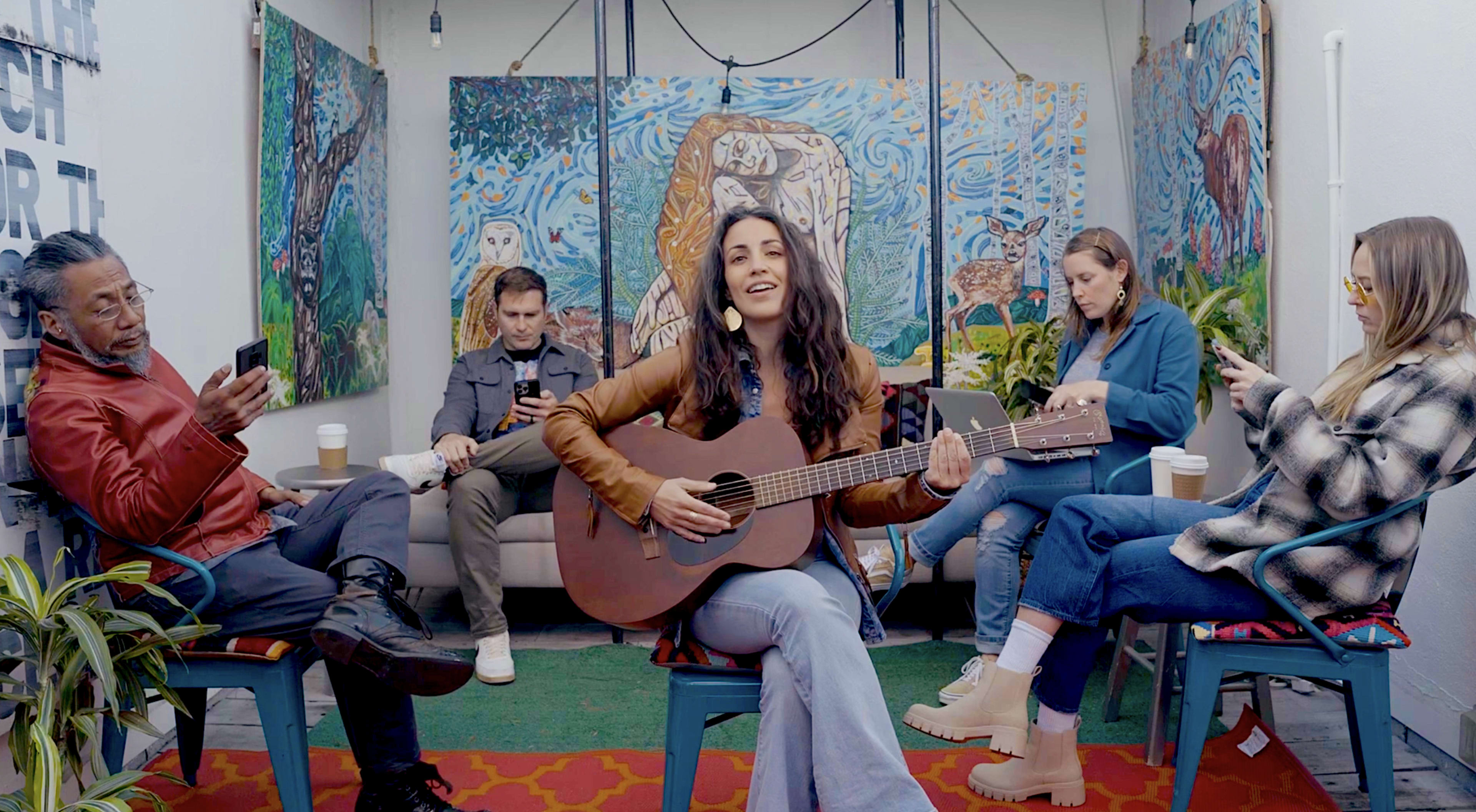 A woman plays guitar and sings in a colorful room while four other people sit nearby, engaged with their phones.
