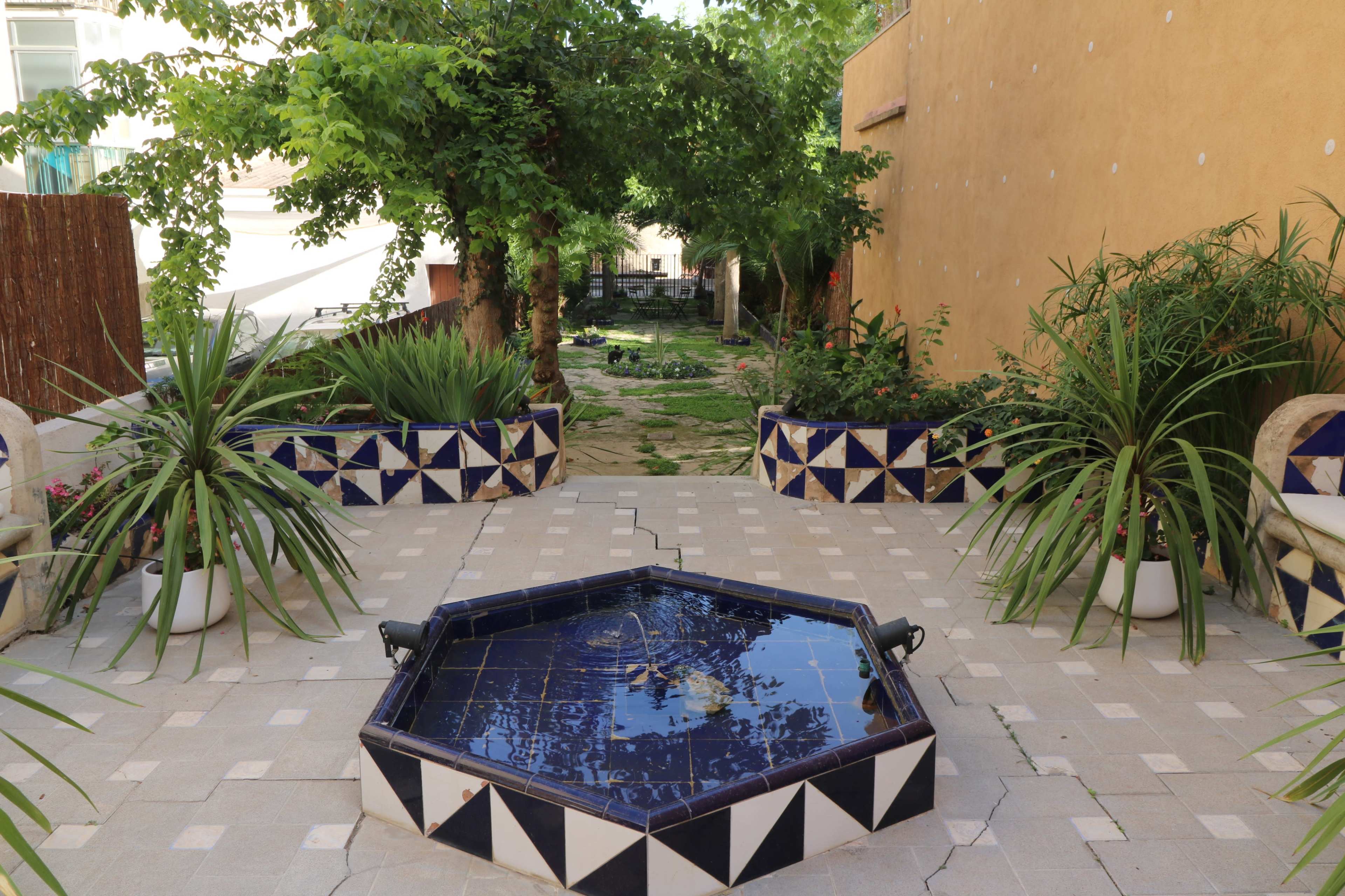 A hexagonal fountain is surrounded by decorative tiled benches and potted plants in a landscaped garden area.