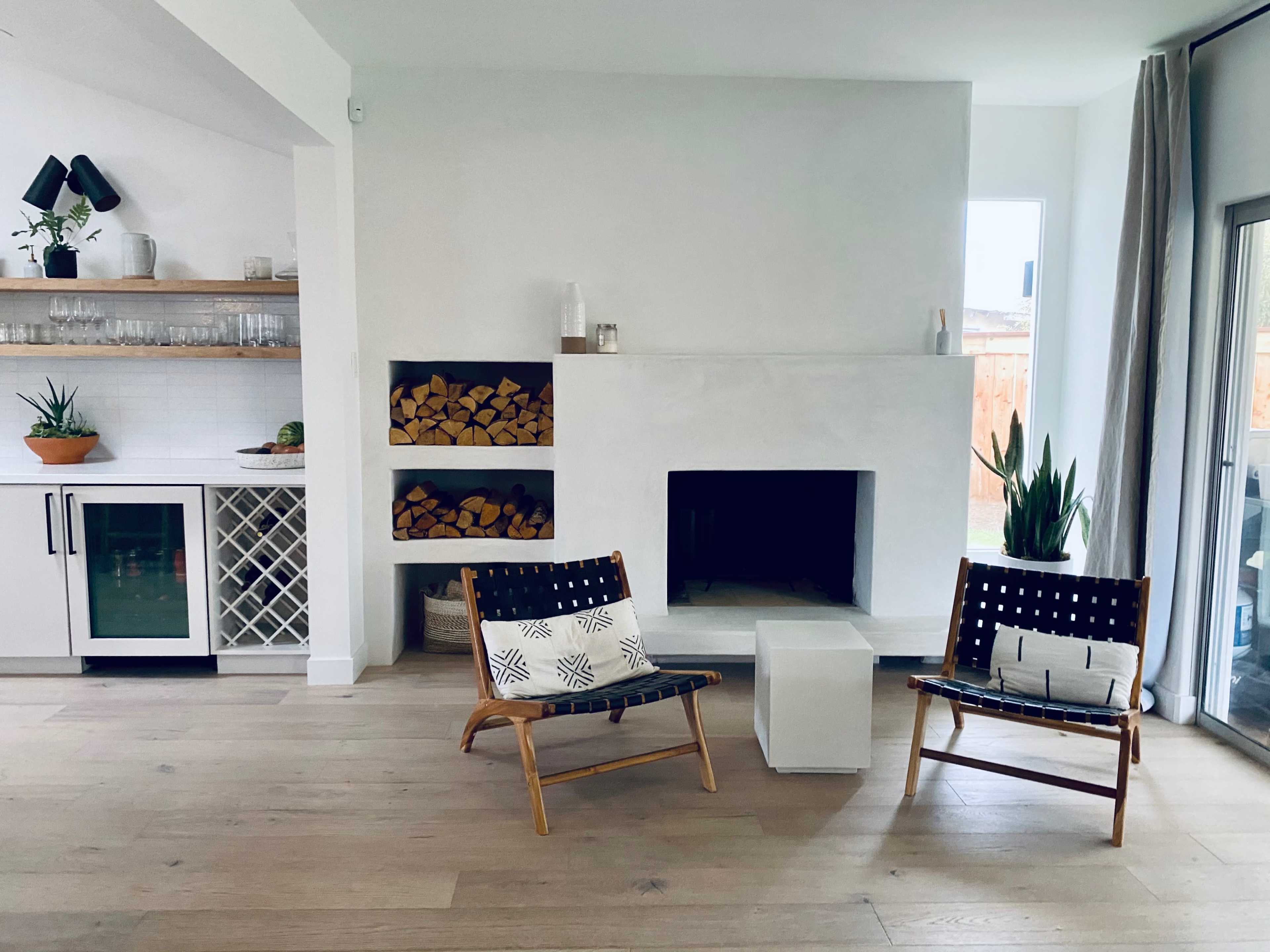 The image shows a modern living room with two woven lounge chairs, a white fireplace, a minimalist coffee table, and a built-in shelf displaying plants and stacked firewood.