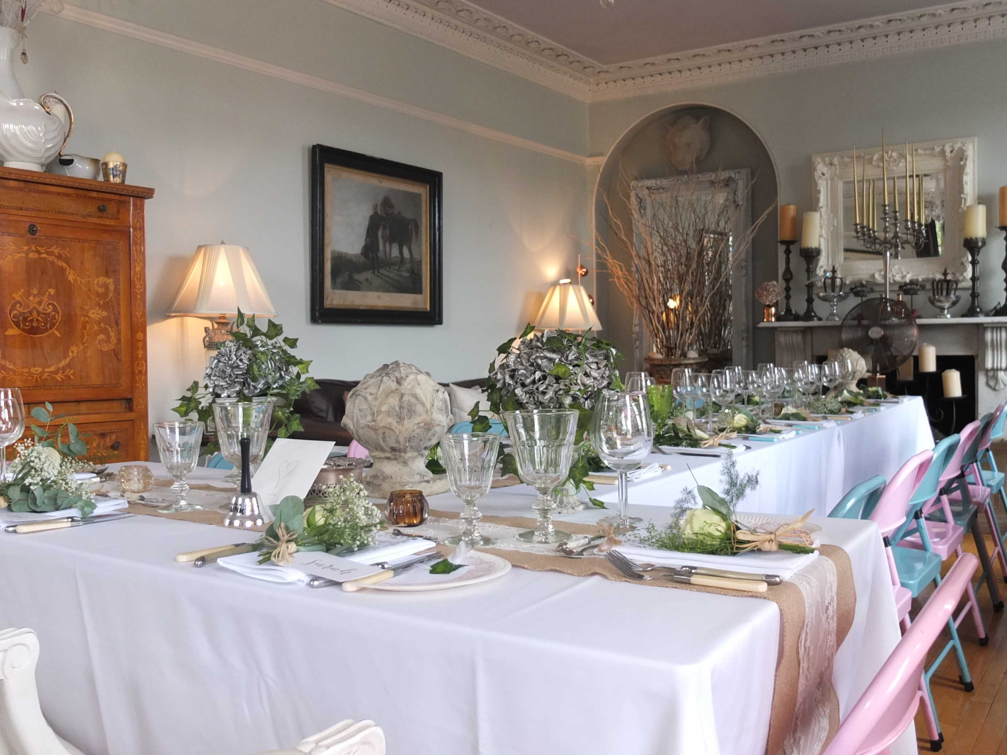 A long banquet table is elegantly set with glassware, cutlery, and decorative elements, surrounded by chairs in a softly lit dining room.