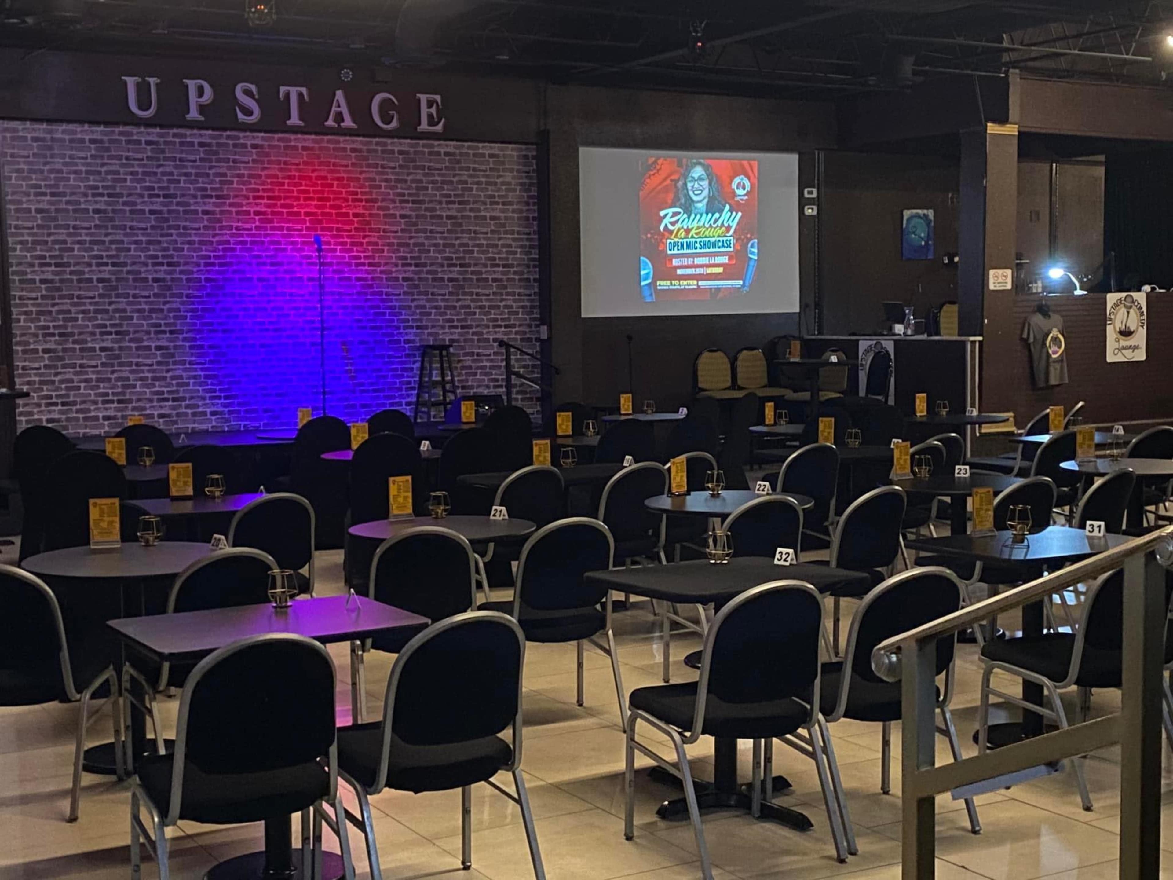 The image shows a performance space labeled "UPSTAGE" with a brick wall backdrop, multiple tables and chairs arranged for an audience, and a large screen displaying an event poster.