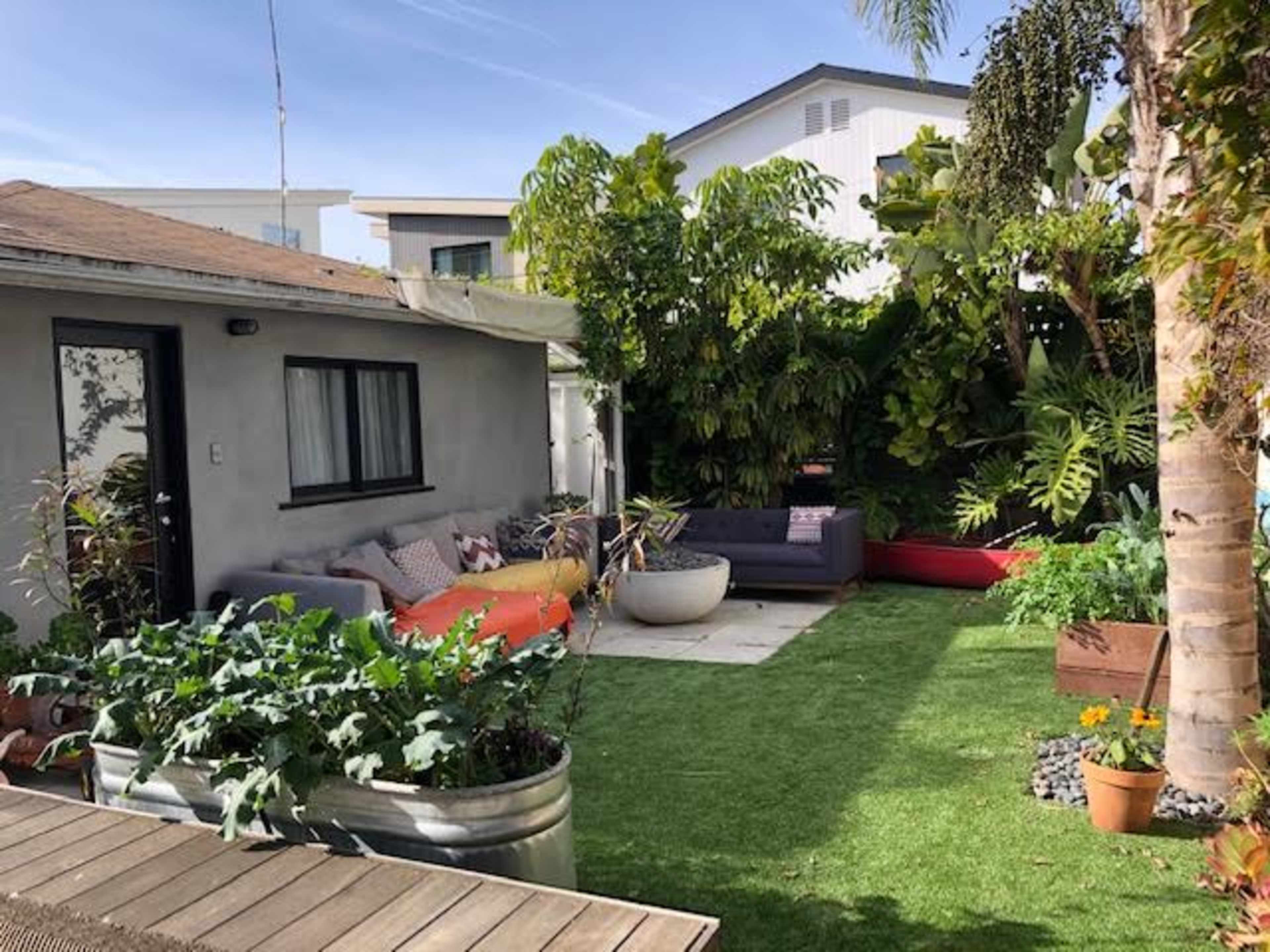 The image shows a garden area featuring a small seating arrangement beside a building, surrounded by various plants and landscaped grass.