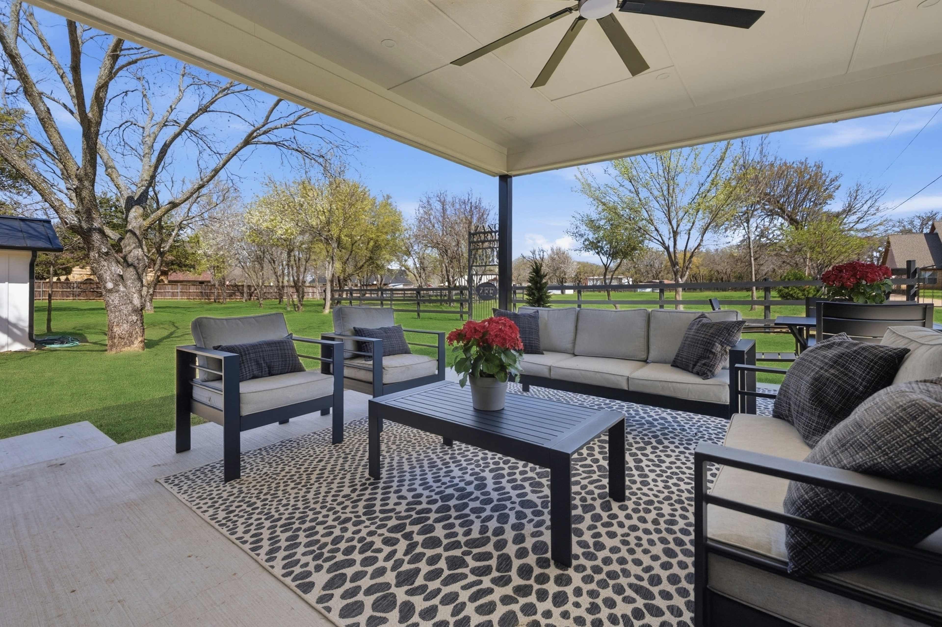 A covered patio features a seating area with gray couches and a coffee table, surrounded by greenery and trees in the background.