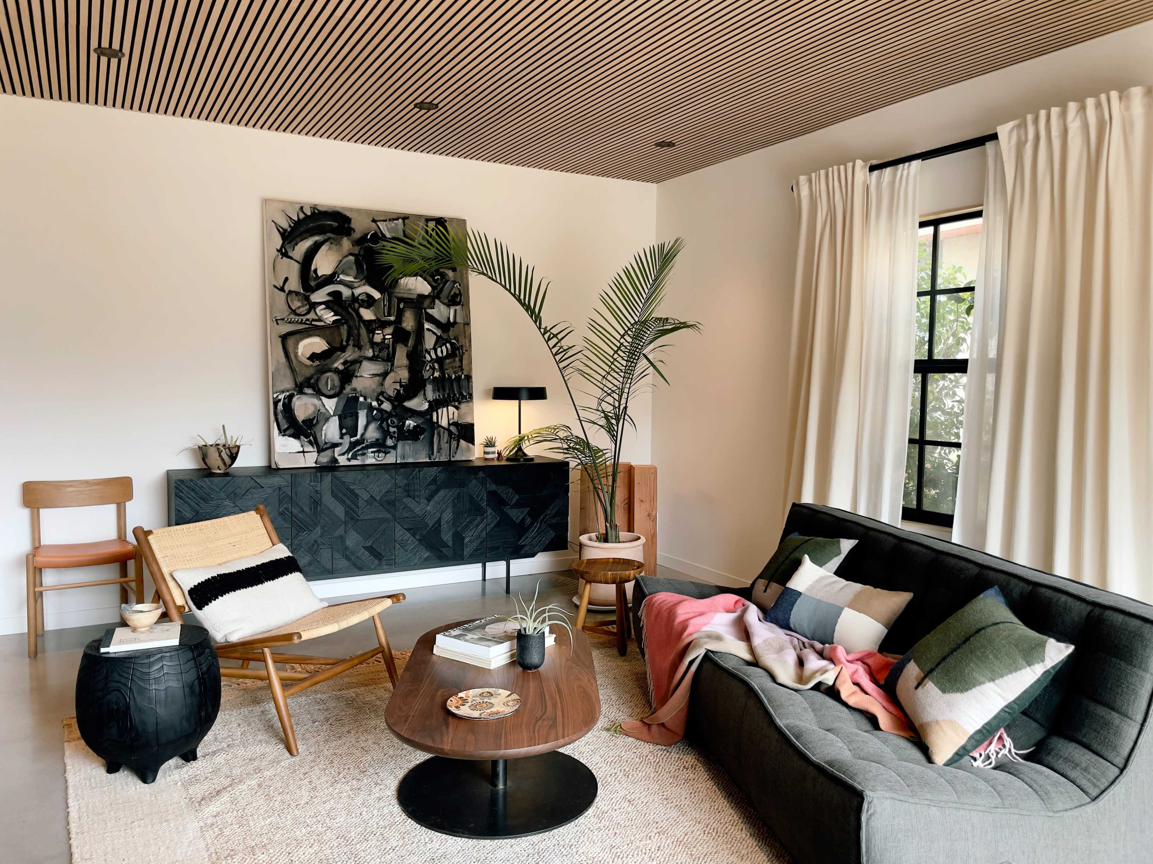 A modern living room with a gray sofa, a round wooden coffee table, a woven rug, a minimalist sideboard, and two chairs, adorned with plants and decorative items.