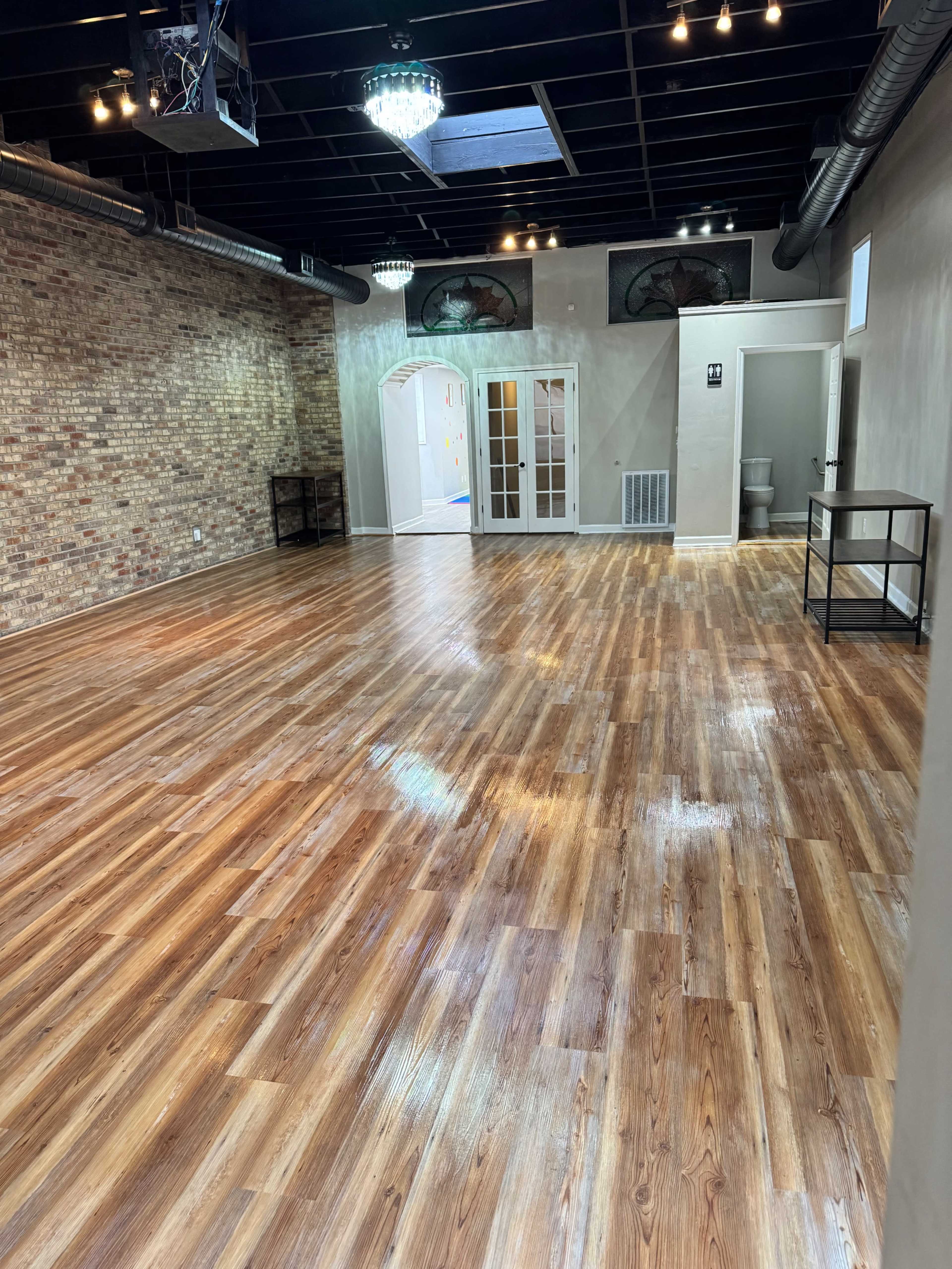 The image shows a spacious, well-lit interior room with a polished wooden floor and brick walls, featuring large windows and doors leading to another area.