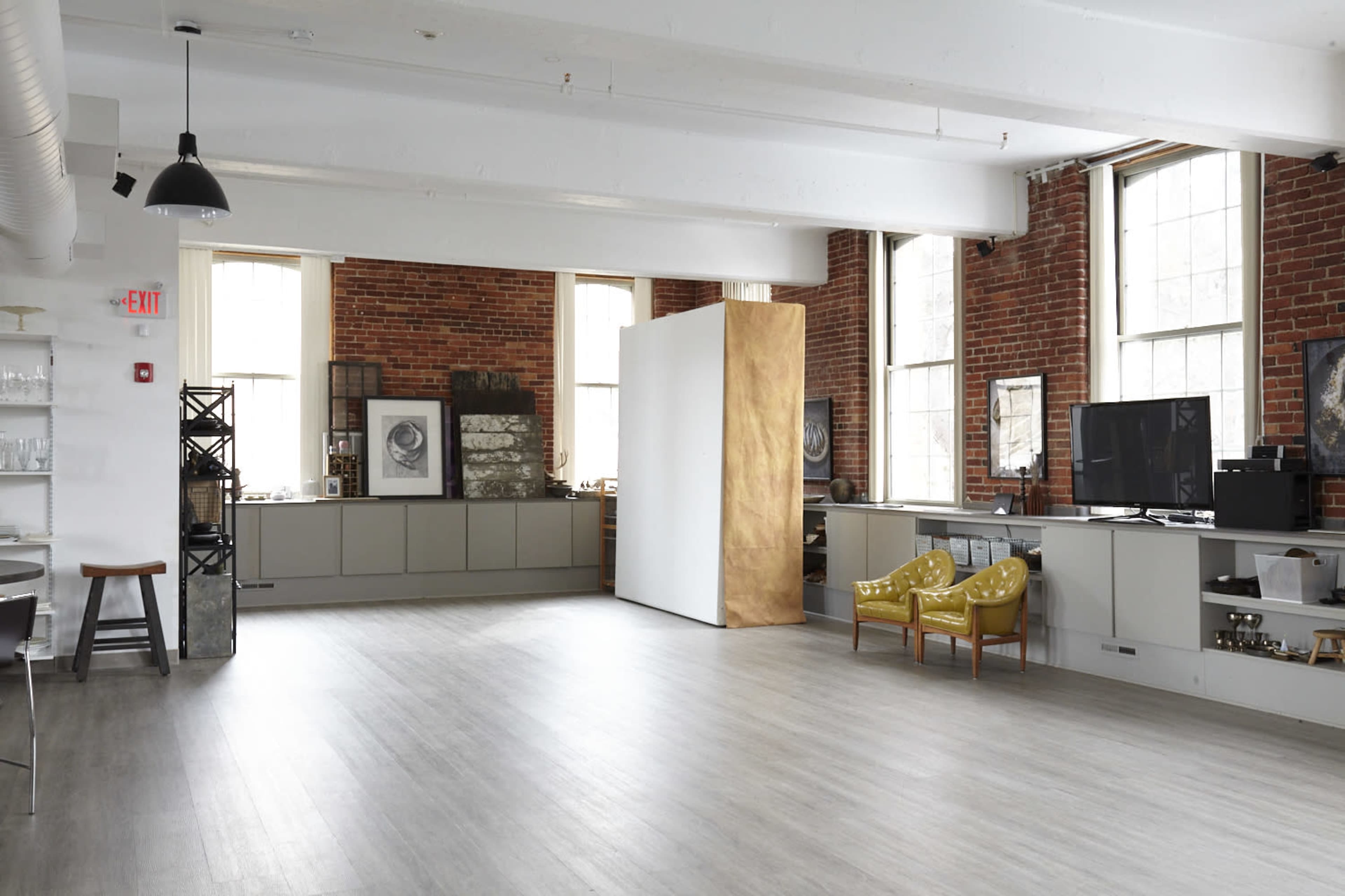 The image shows a spacious, industrial-style room with large windows, brick walls, and a modern decor featuring a gold partition and yellow chairs.