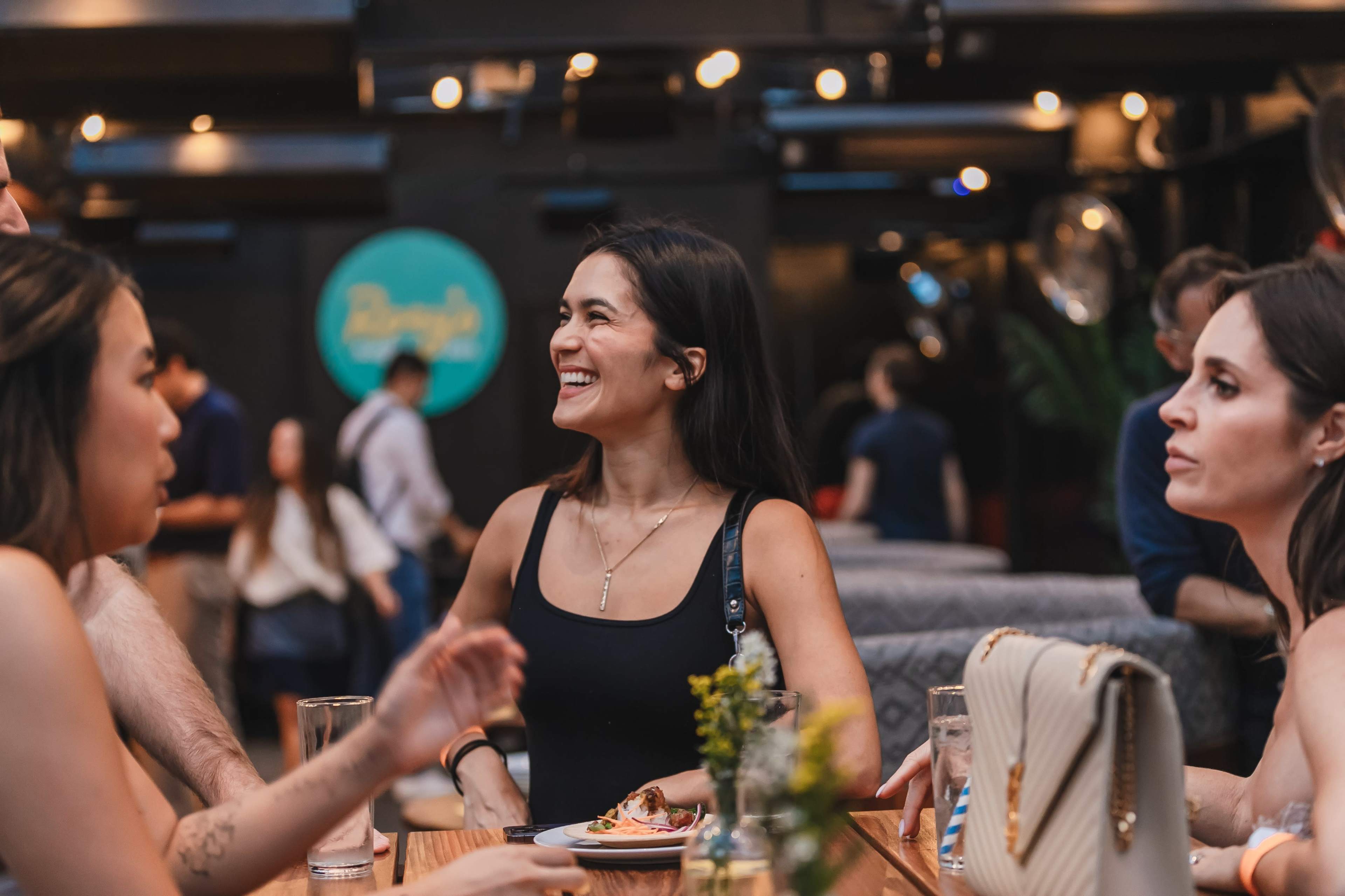 A group of people is gathered around a table at a lively indoor venue, with one woman laughing while others engage in conversation.
