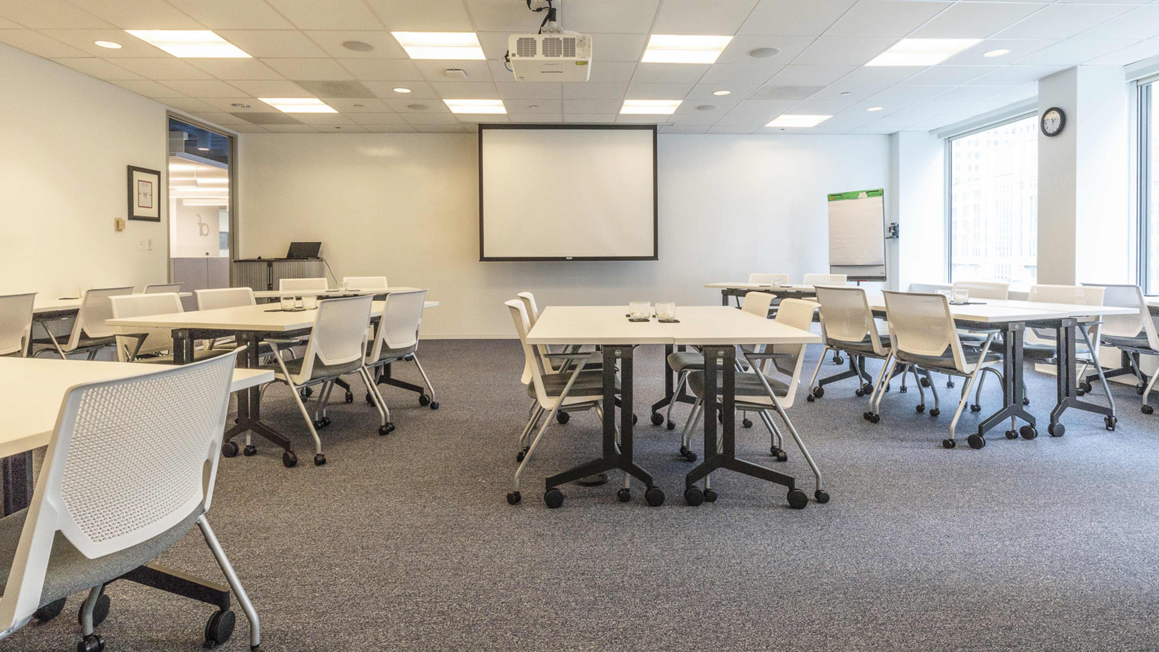 A spacious meeting room features several white tables and chairs arranged for a presentation, with a projector screen at the front and windows providing natural light.