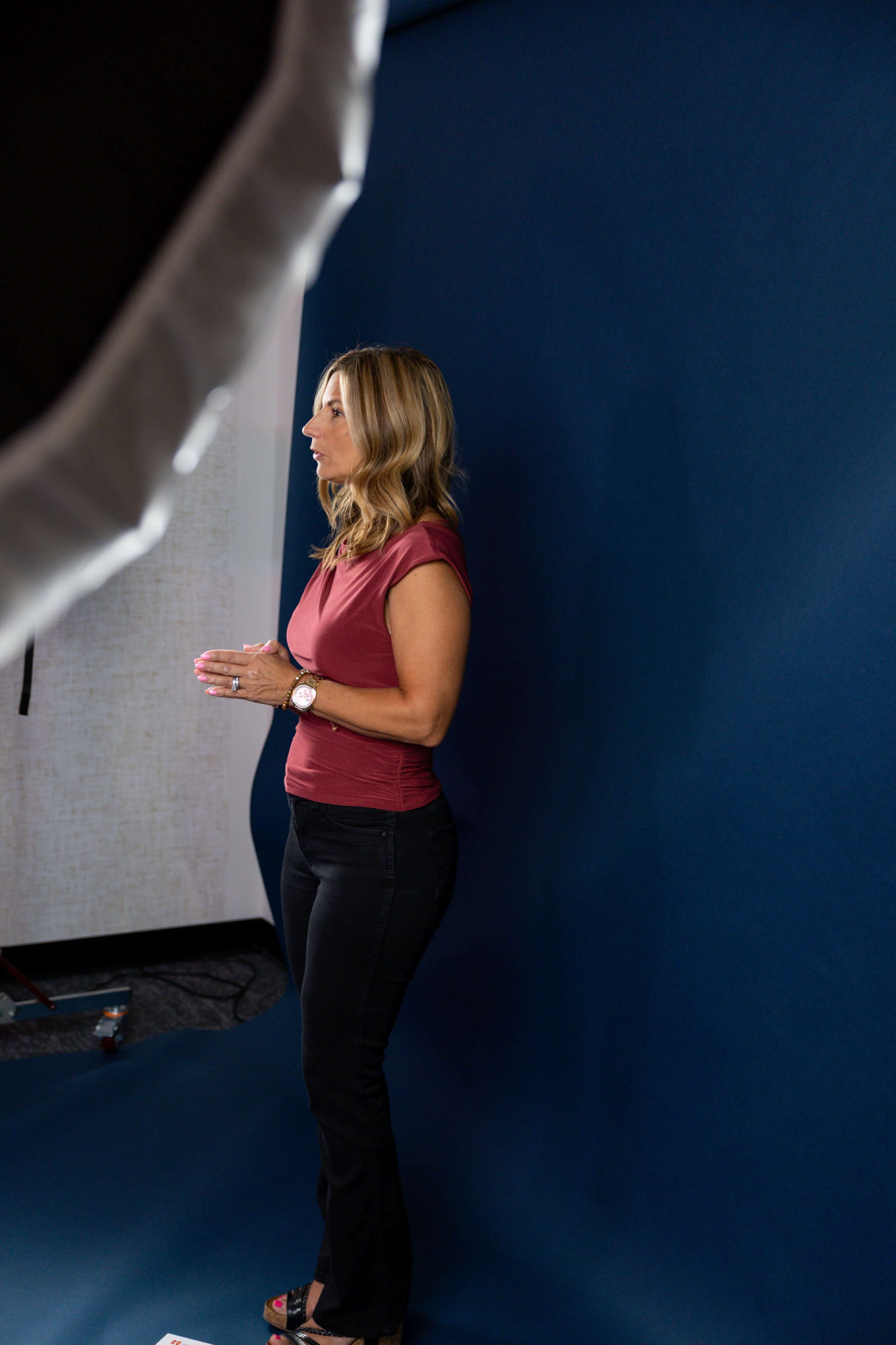 A woman stands on a blue backdrop, gesturing as she speaks.
