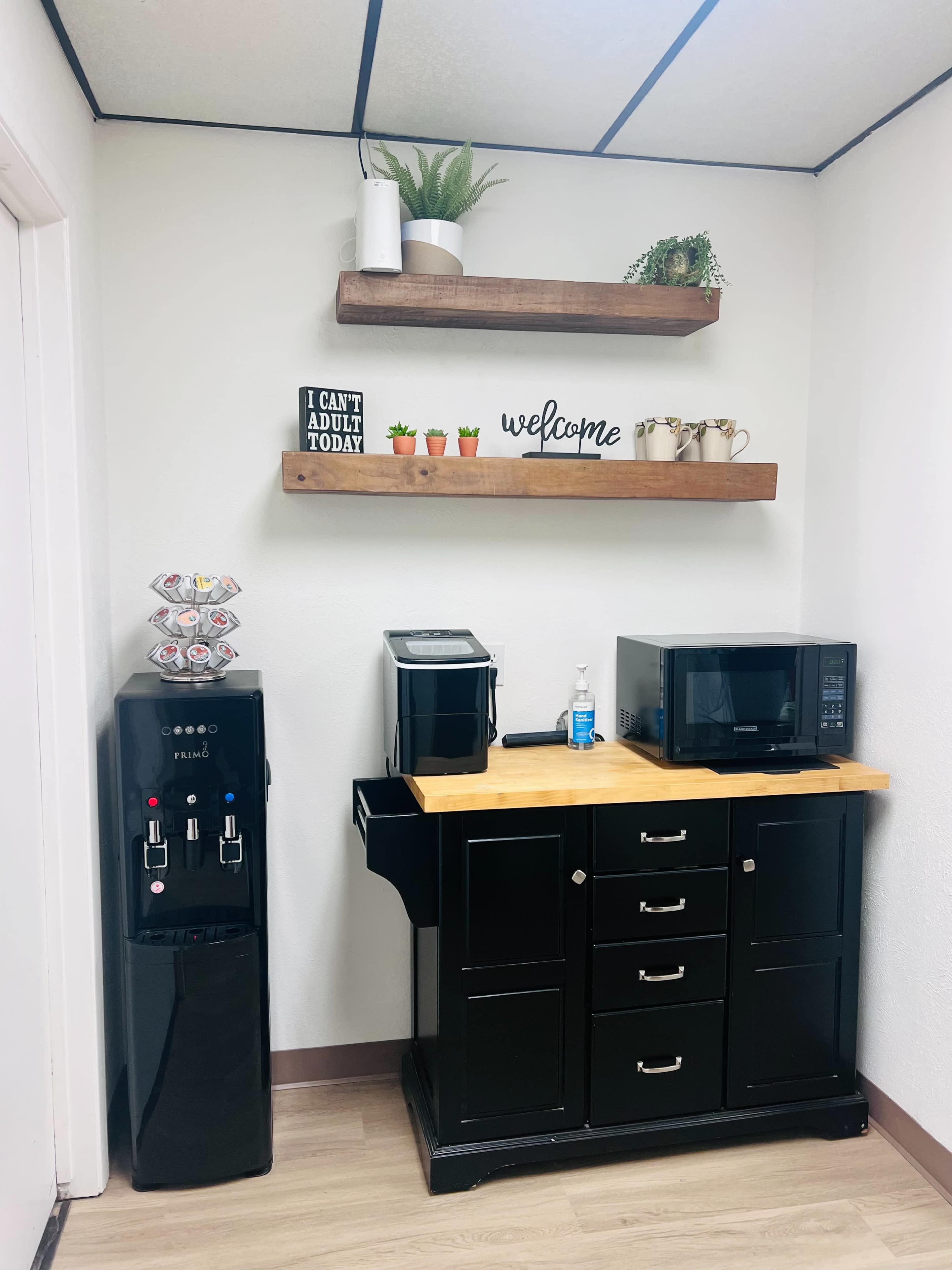 A small break area with a black water cooler, a coffee maker, a microwave, and a wooden countertop with drawers, alongside wall-mounted shelves displaying plants and decorative items.