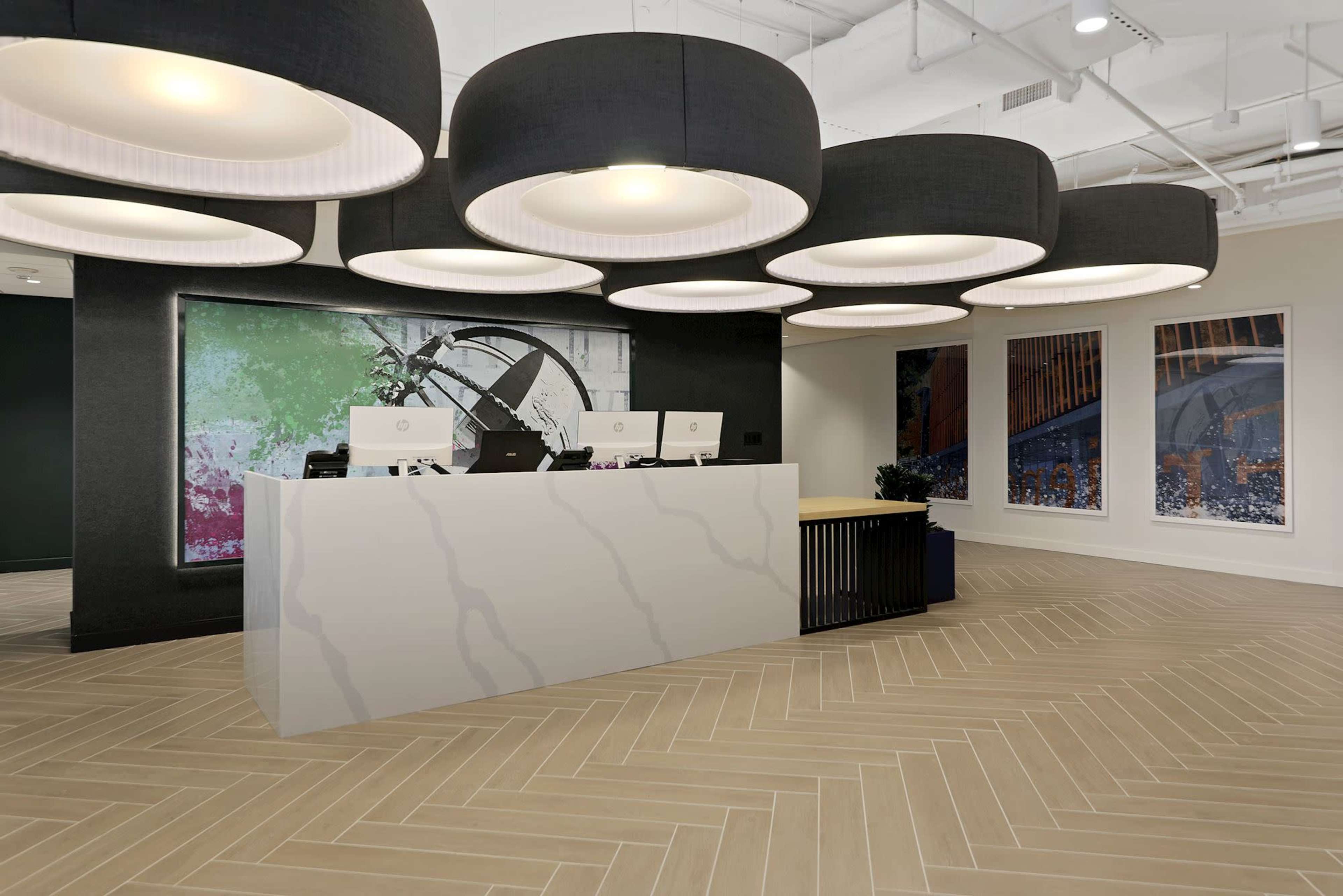 A modern reception area features a white marble front desk with multiple computer stations, surrounded by large black pendant lights and colorful abstract artwork on the walls.