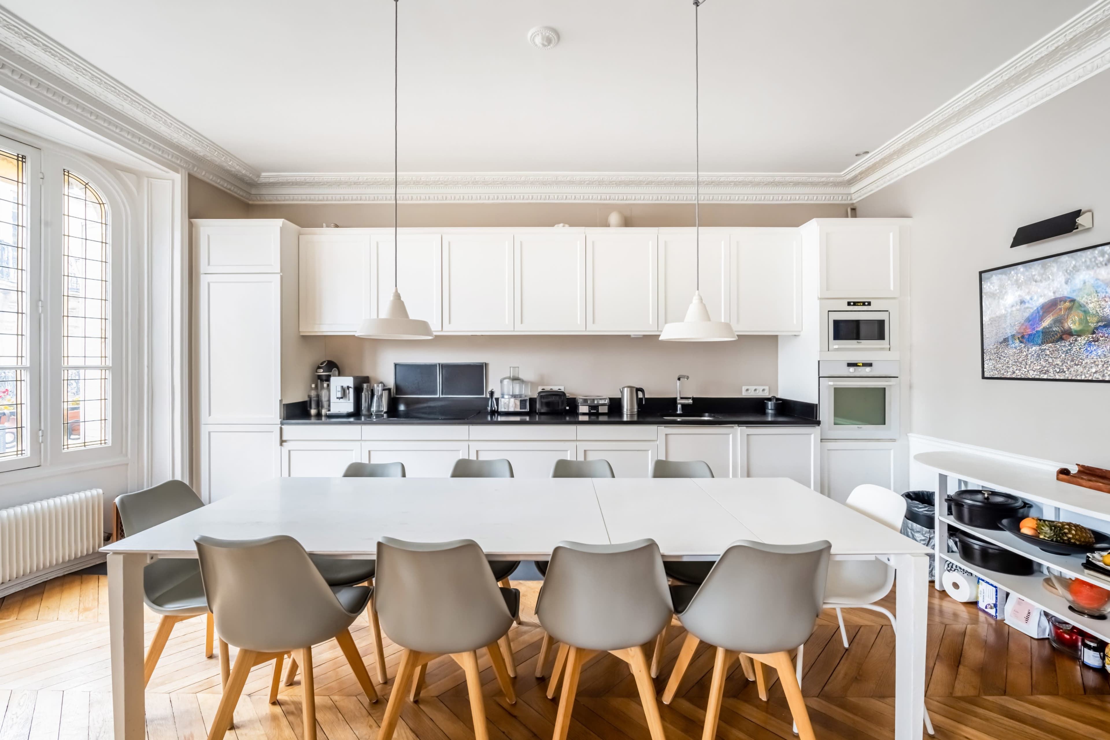 The image depicts a modern kitchen with a large white table surrounded by gray chairs, featuring white cabinets and dark countertops.
