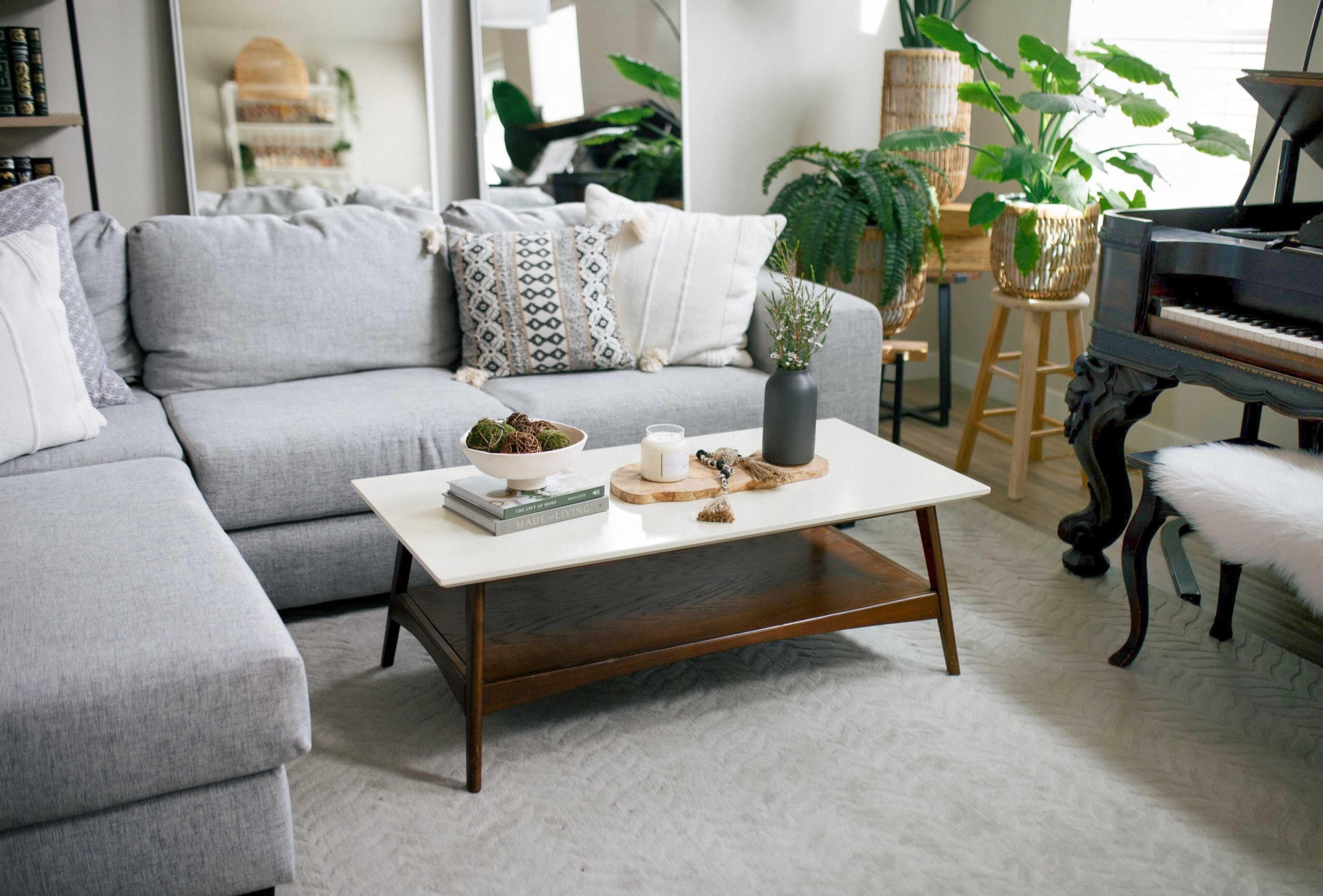 A modern living room features a light gray sectional sofa, a wooden coffee table with decorative items, plants in the background, and a piano nearby.