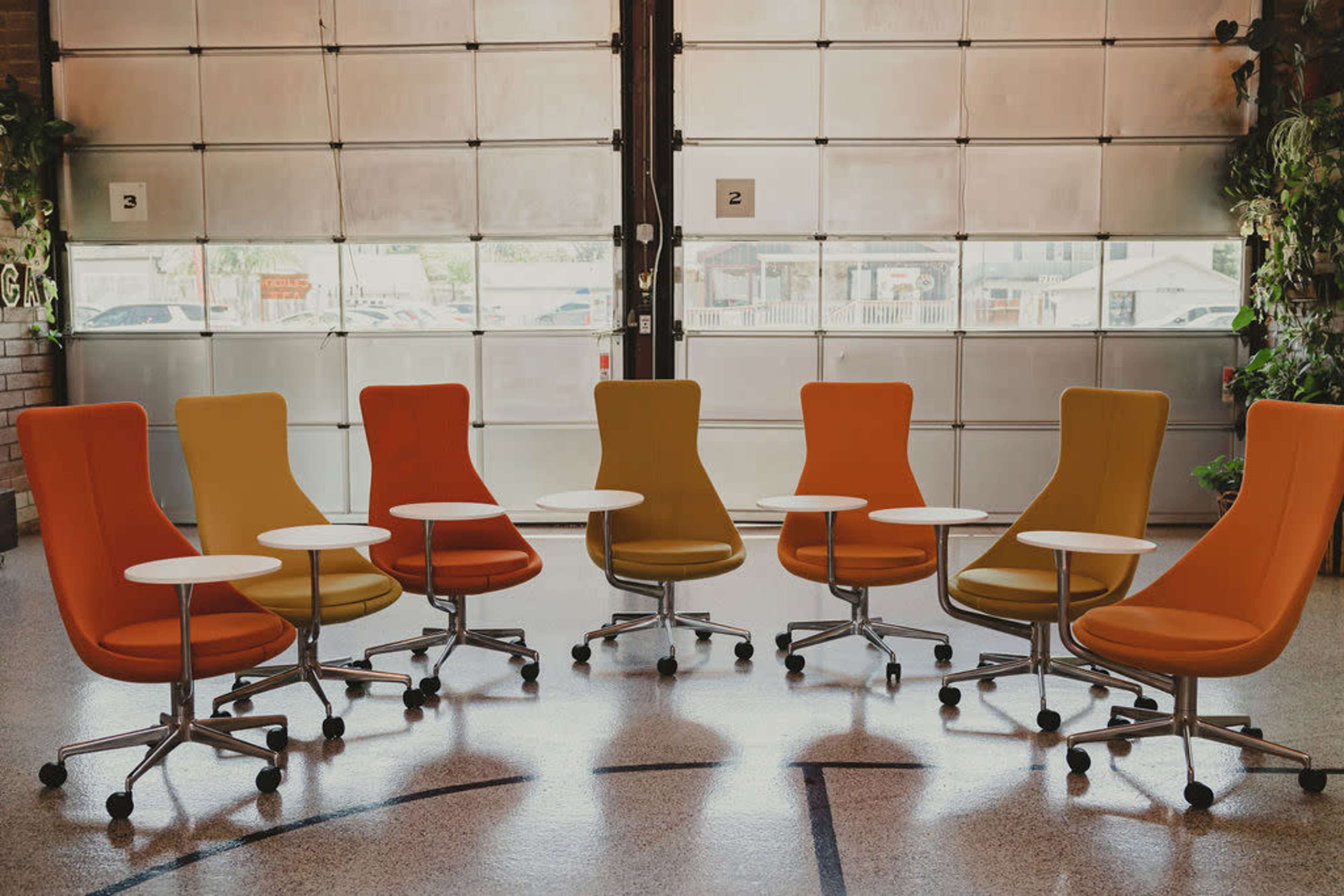 A group of five modern swivel chairs in yellow, orange, and gray is arranged around small white tables in a spacious room with large windows.