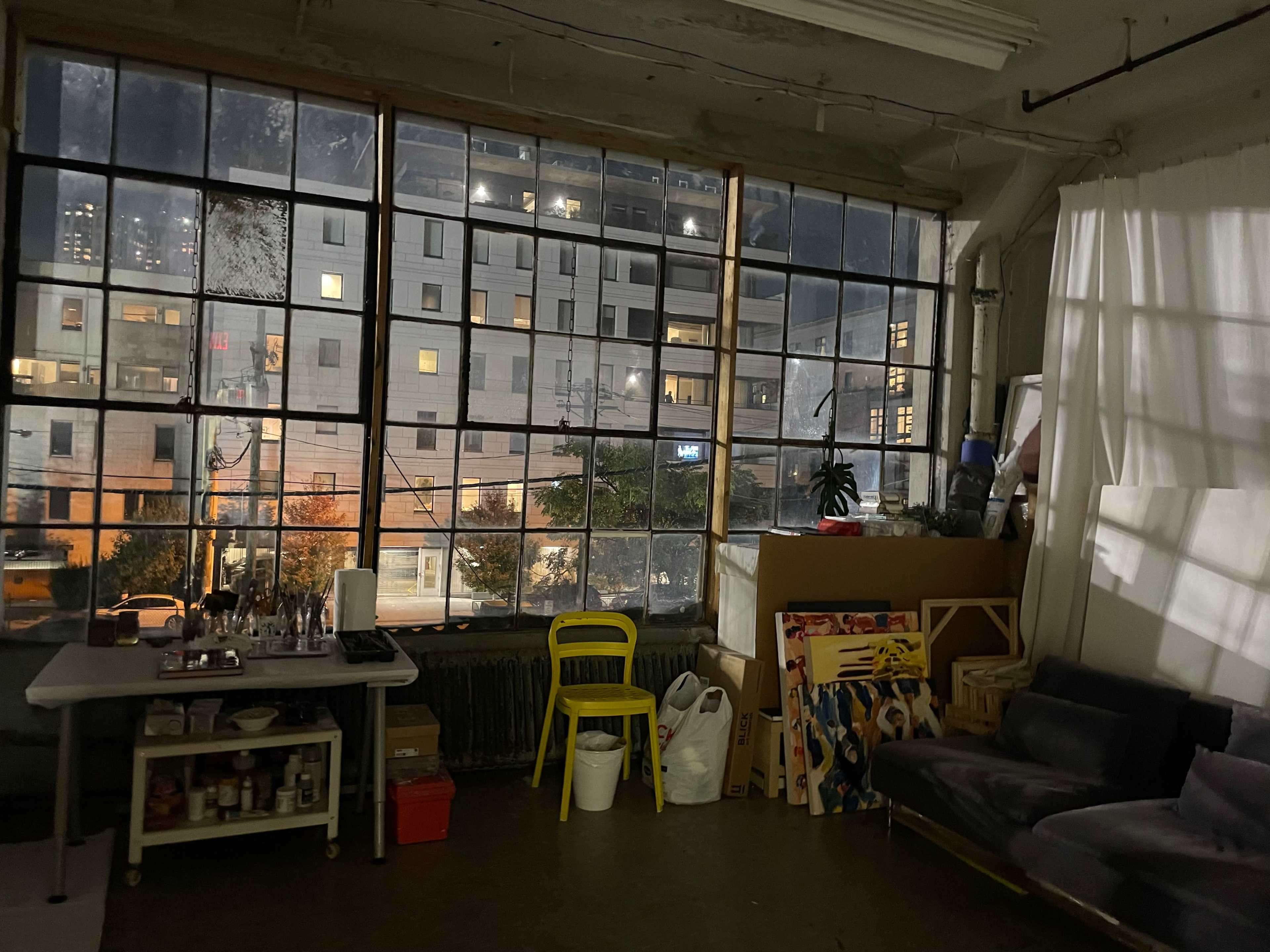 The image shows a large window with multiple panes, revealing a cityscape at night, alongside a cluttered room featuring a table, a chair, and various boxes.