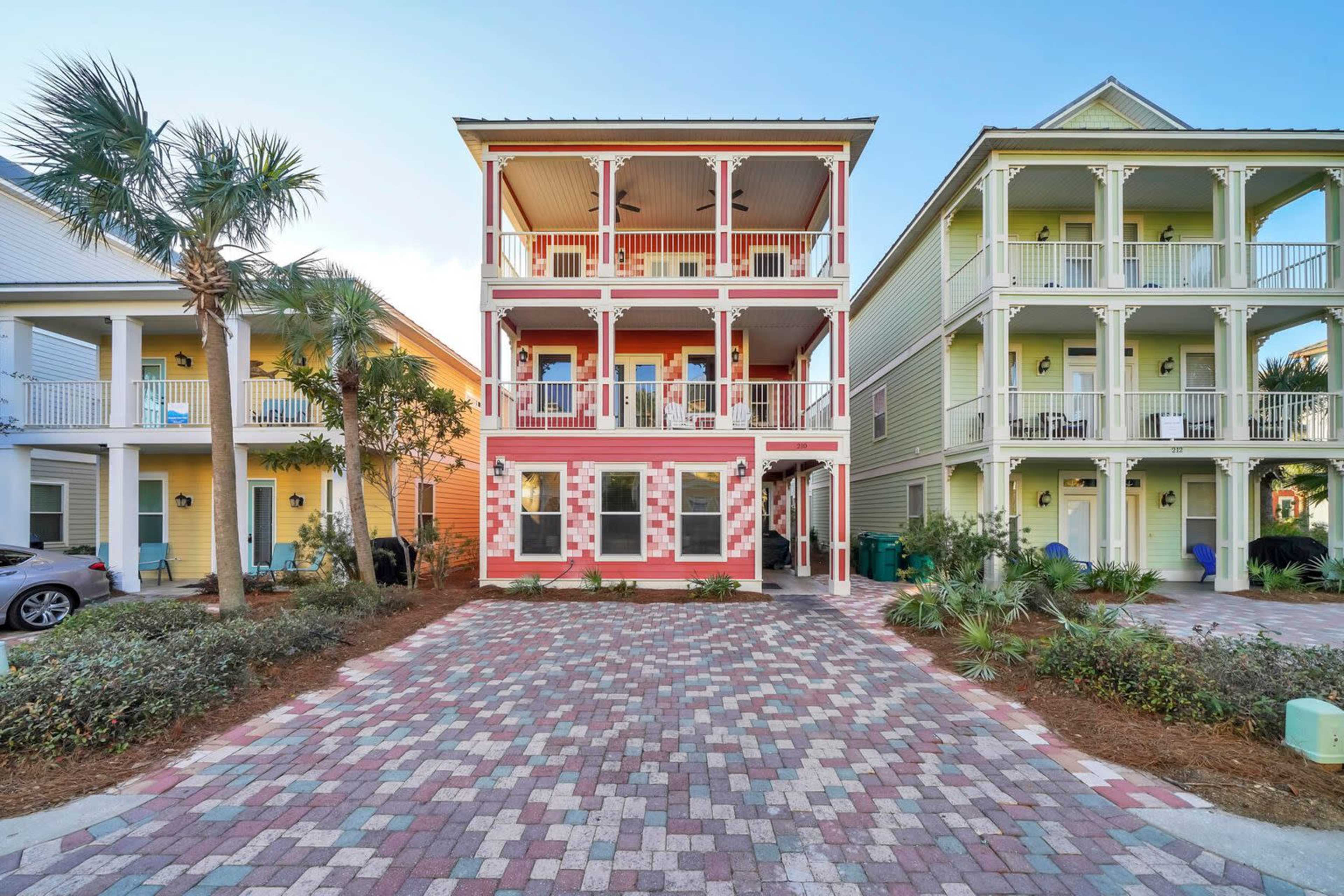 A row of three colorful, multi-story houses with porch balconies, surrounded by landscaped greenery and a paved driveway.