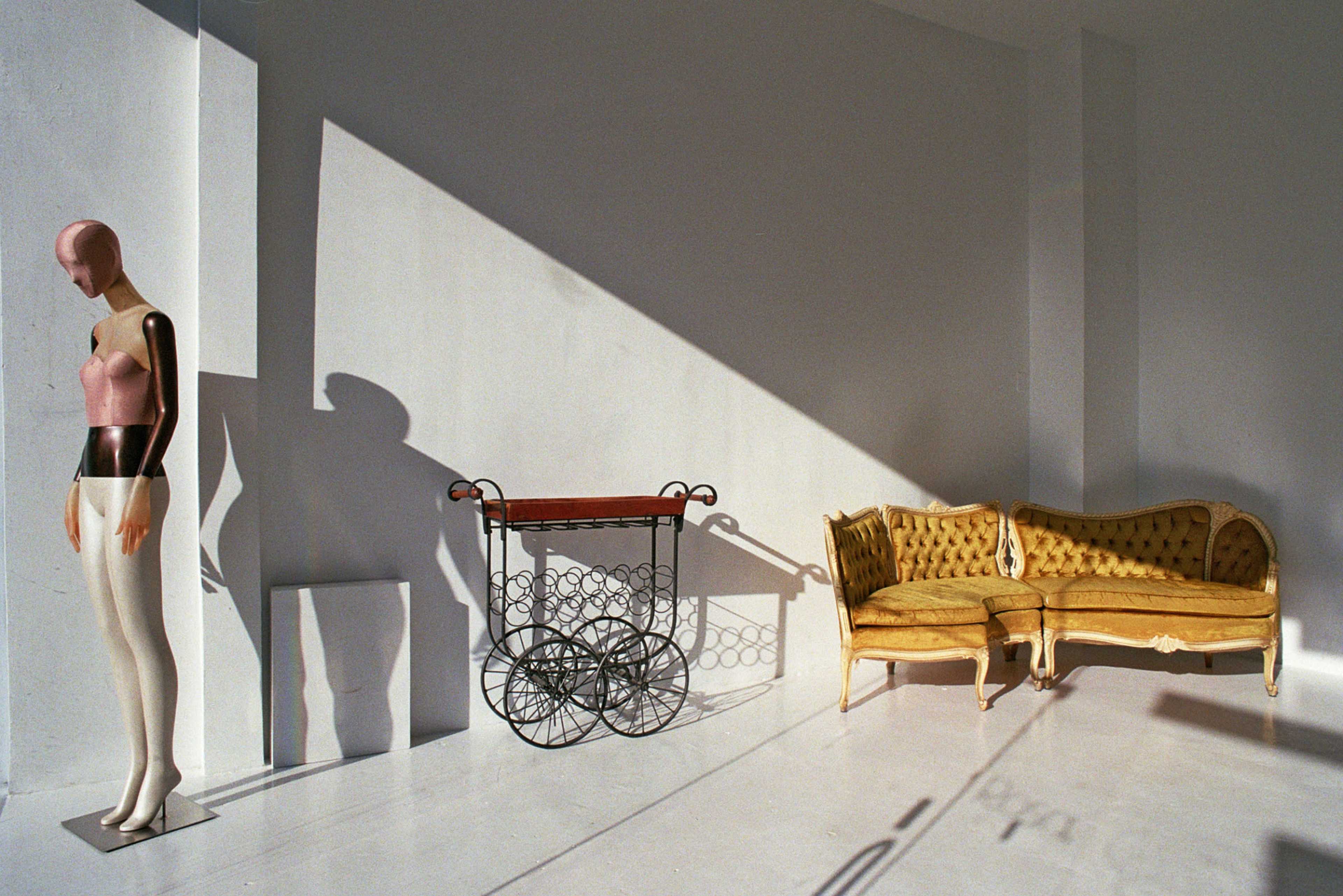 The scene features a mannequin, an ornate cart, and a vintage sofa casting distinct shadows in a well-lit, minimalist room.