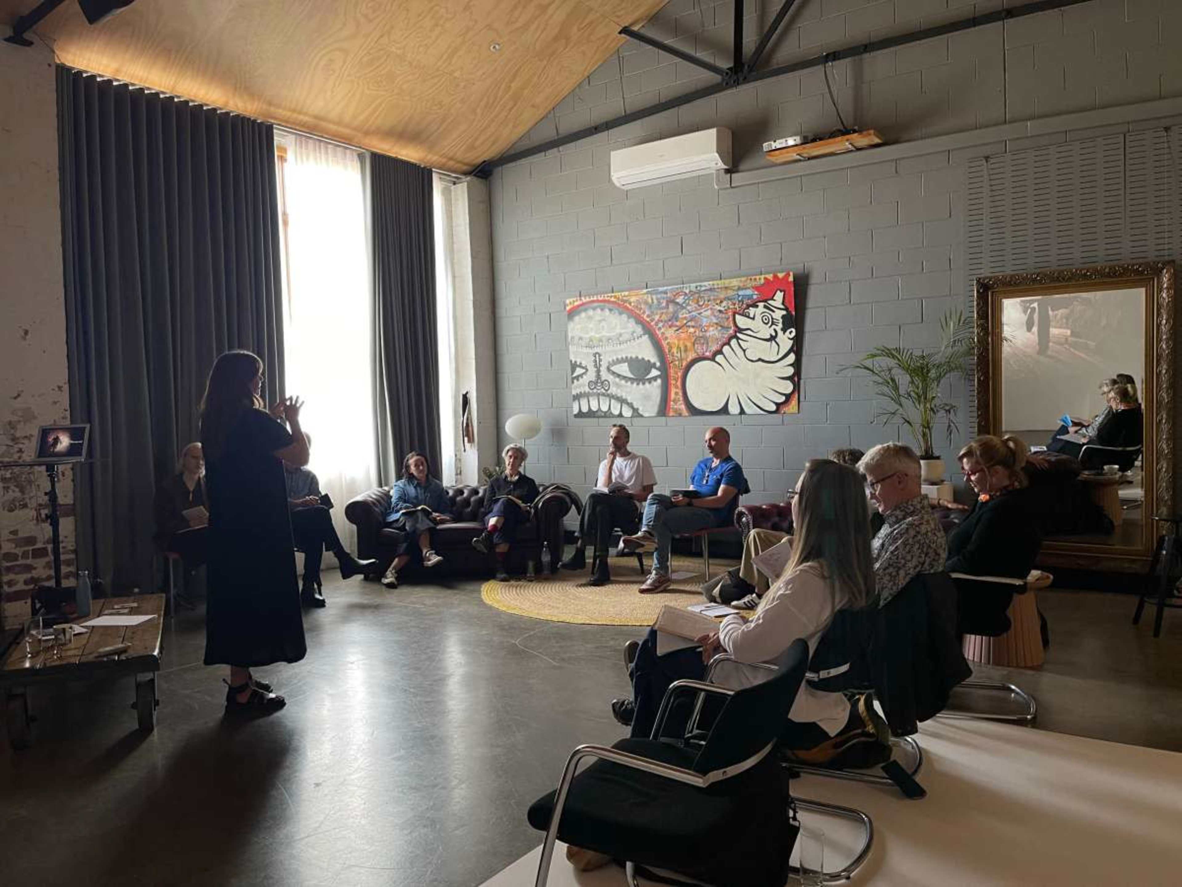 A group of people is seated in a spacious room while a woman stands and speaks in front of them.