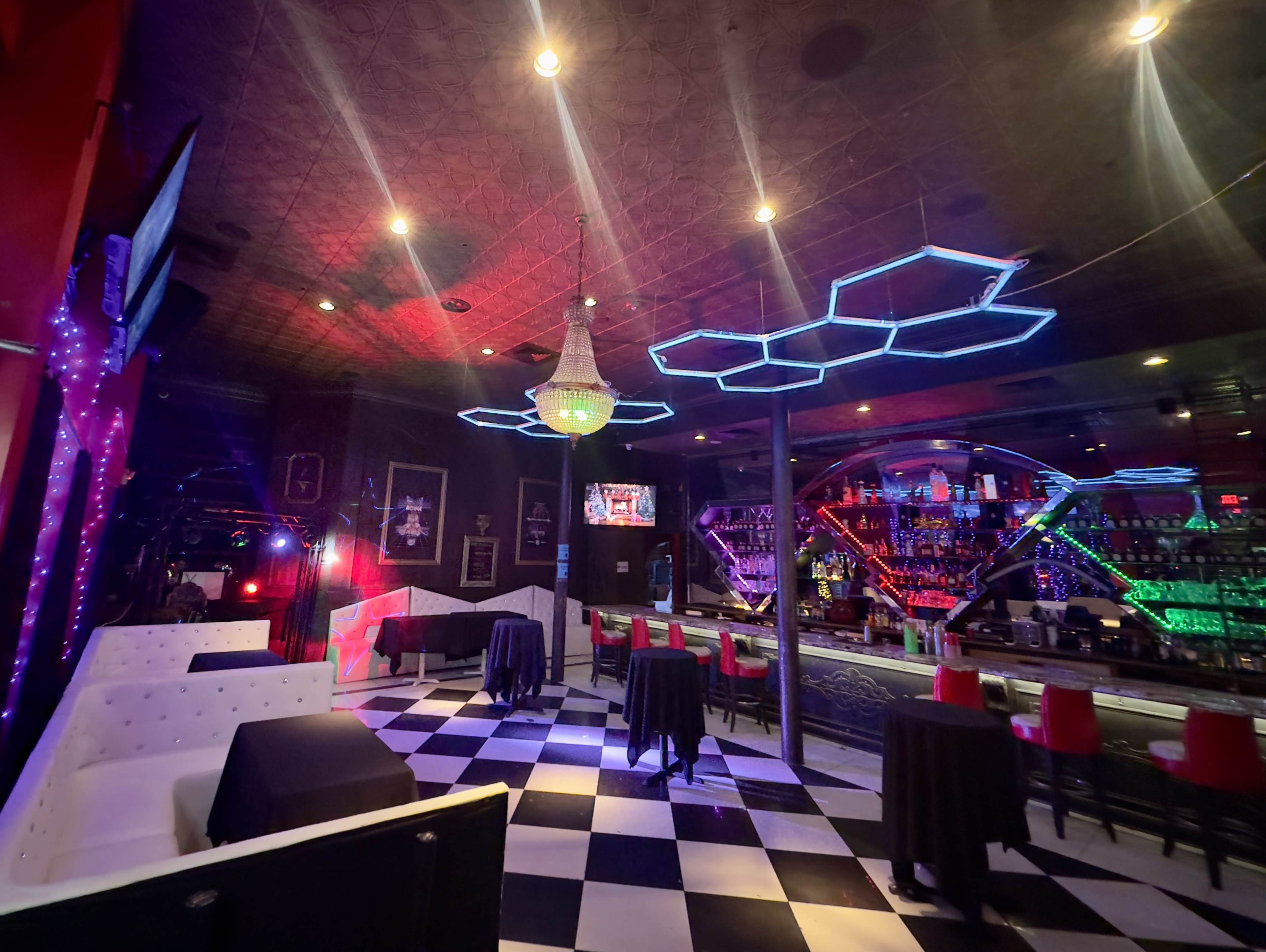 The image shows a dimly lit bar with black and white checkered flooring, modern furniture, and colorful LED lighting illuminating the space.