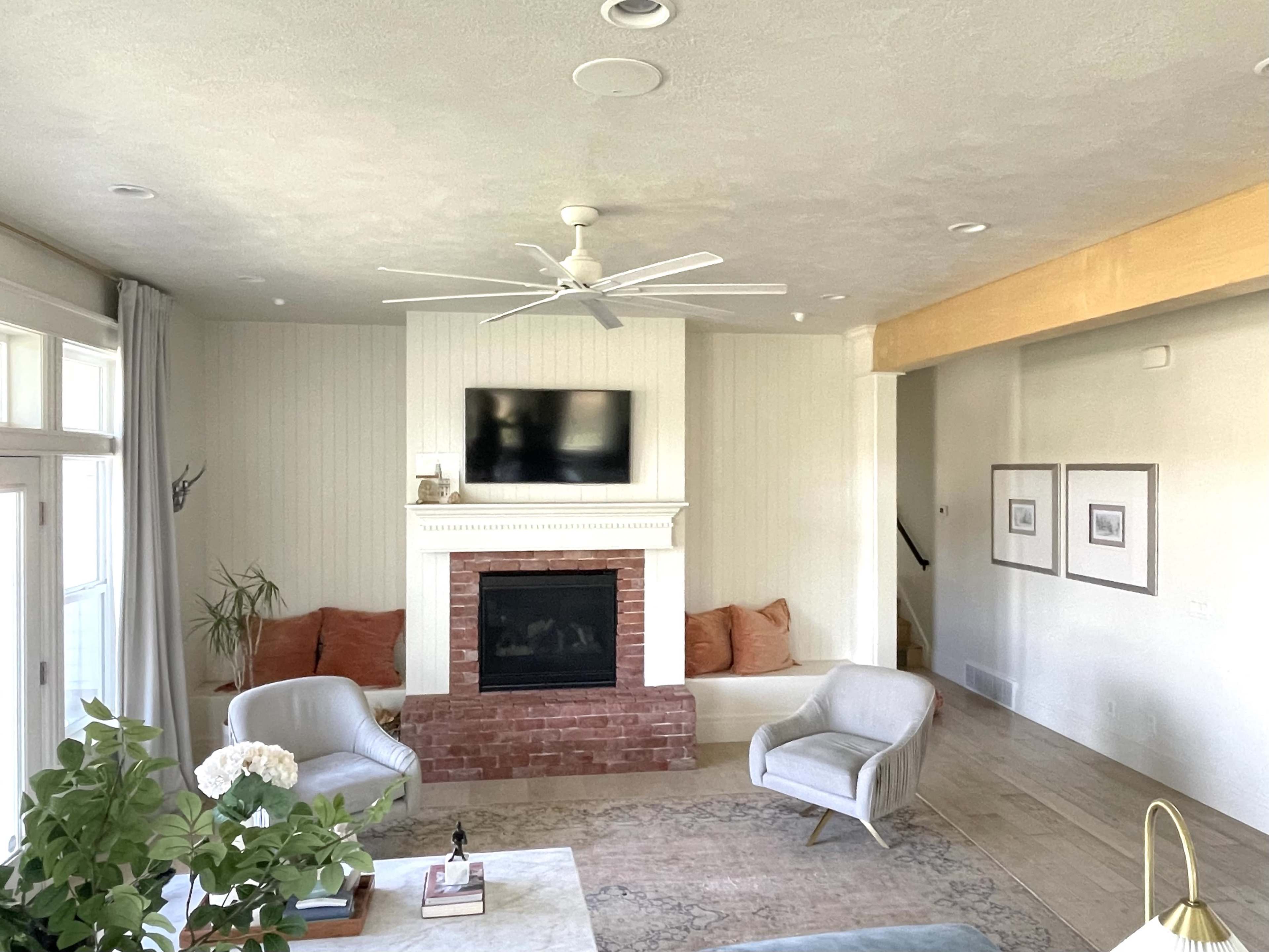 A spacious living room features a brick fireplace, a mounted television, two light-colored chairs, and a sectional sofa with orange cushions.