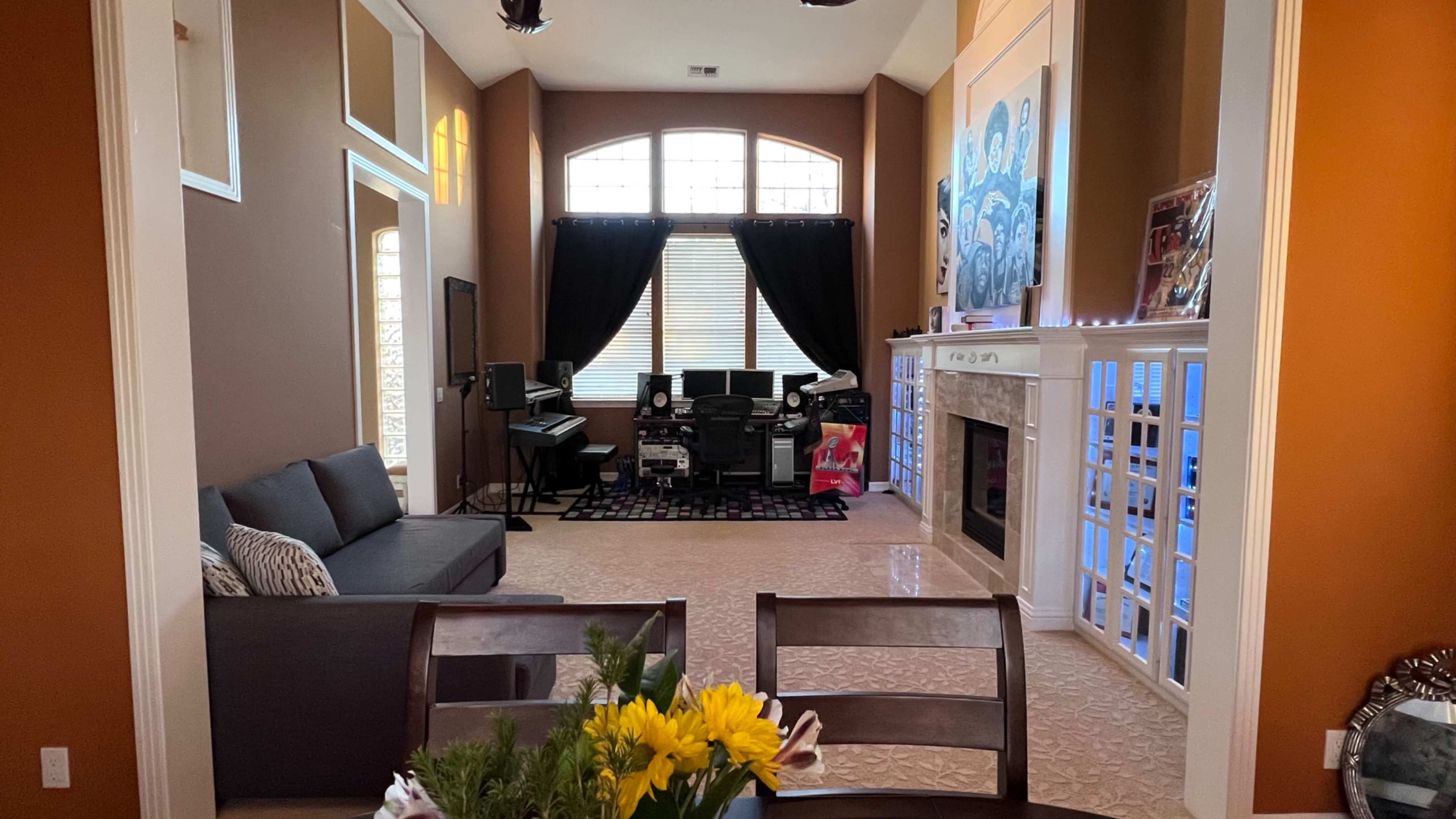 A living room with a sofa, a window with black curtains, a music setup on the left, and a fireplace with decorative items on the right.