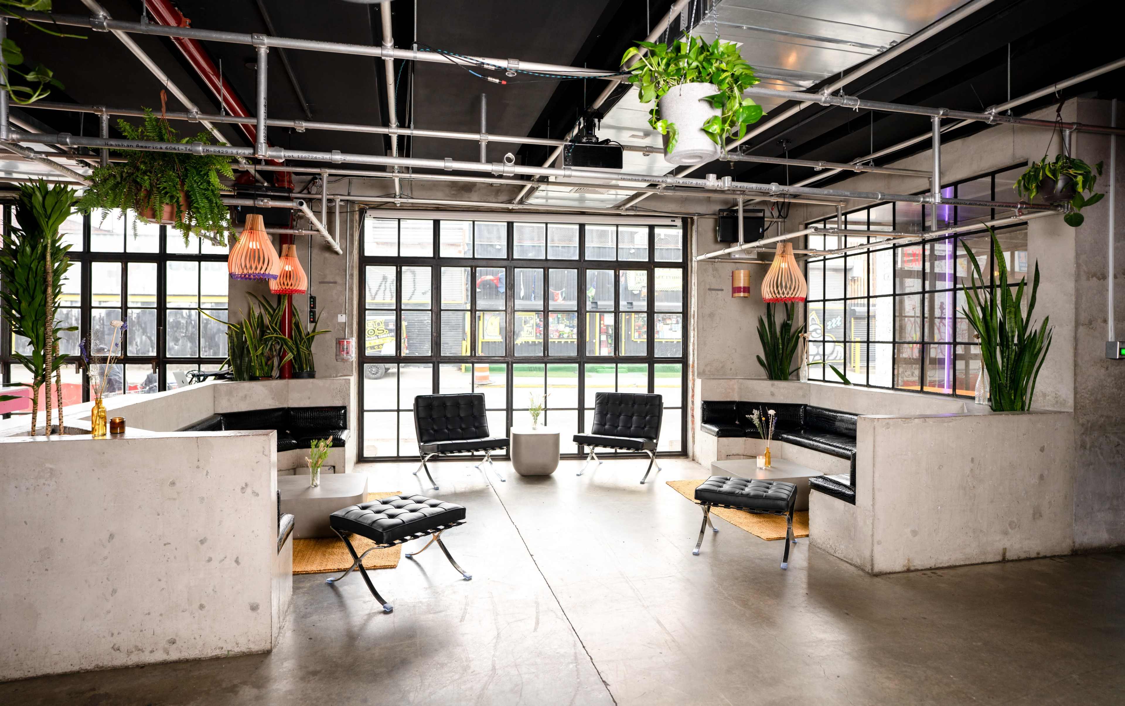 The image shows a modern lounge area with black seating, concrete walls, and large windows, featuring greenery and pendant lights.