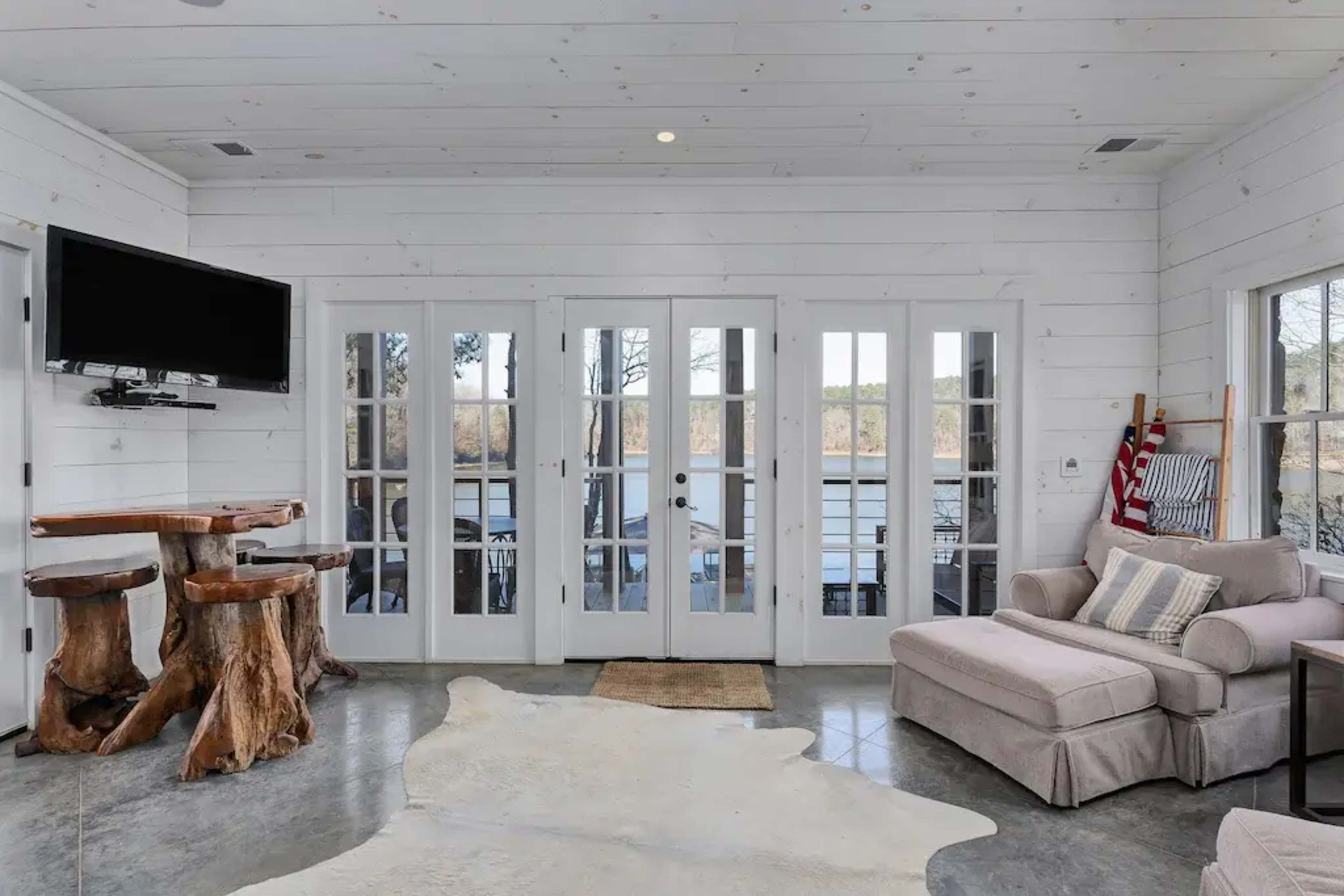 A room with white wooden walls, a seating area featuring a light-colored sofa, a wooden table made from tree stumps, and glass doors leading to an outdoor view.