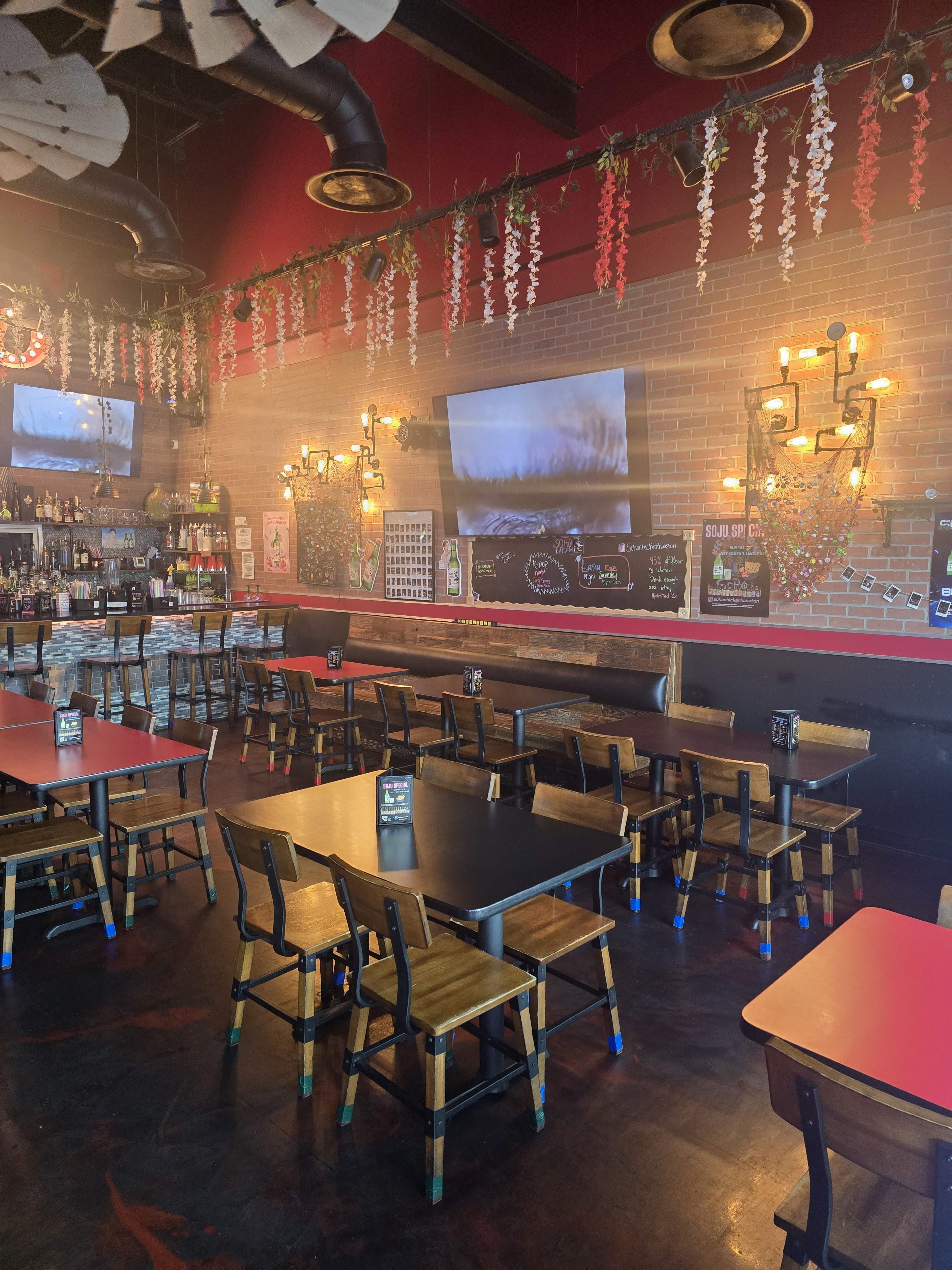 The image shows a restaurant interior with wooden tables and chairs, a bar area in the background, and decorative lighting along the walls.