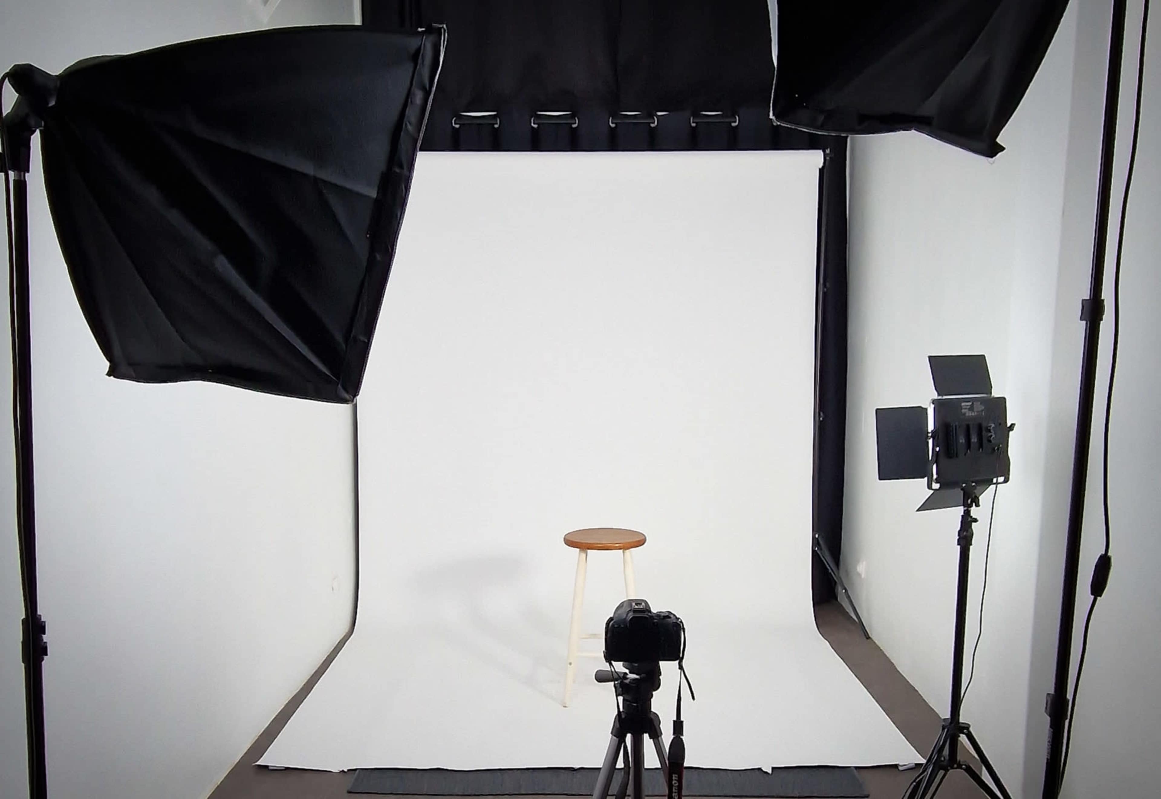 A photography studio setup features a plain white backdrop, a wooden stool, and various lighting equipment positioned around a camera on a tripod.