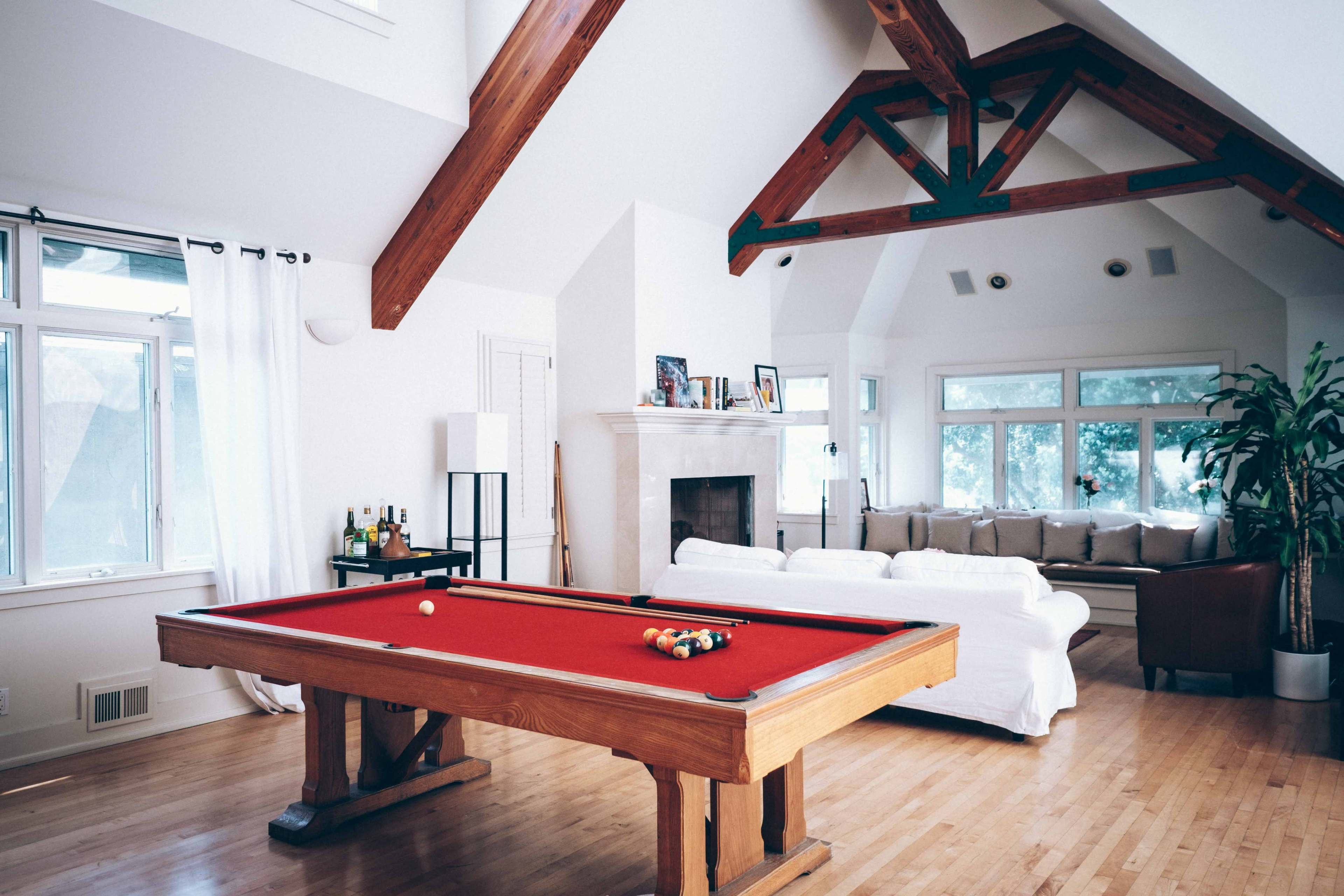 A room features a pool table with a red felt surface, surrounded by large windows and a cozy seating area.