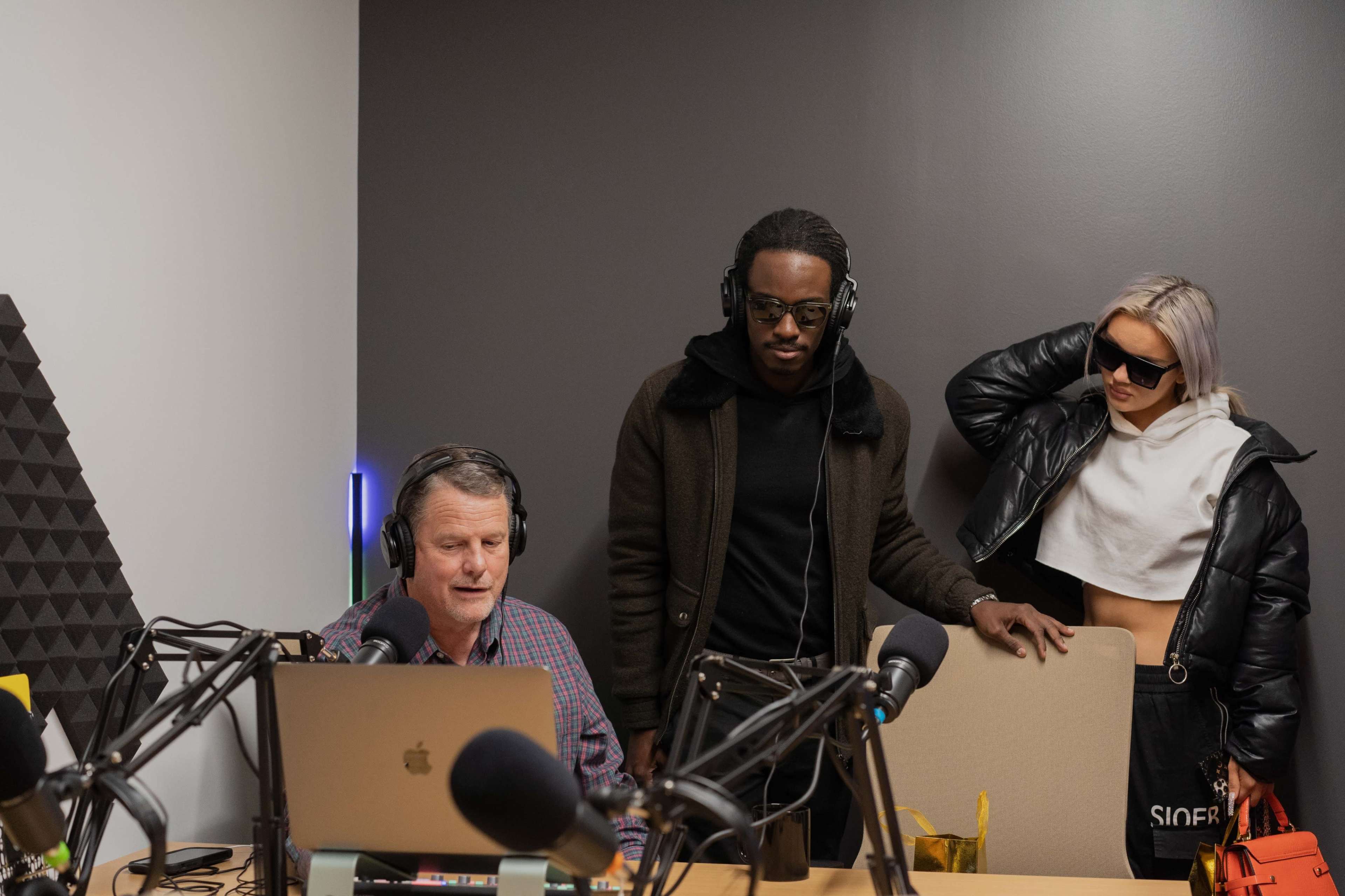 Three individuals are in a recording studio setting, with one person seated at a laptop and two others standing nearby, all wearing headphones.
