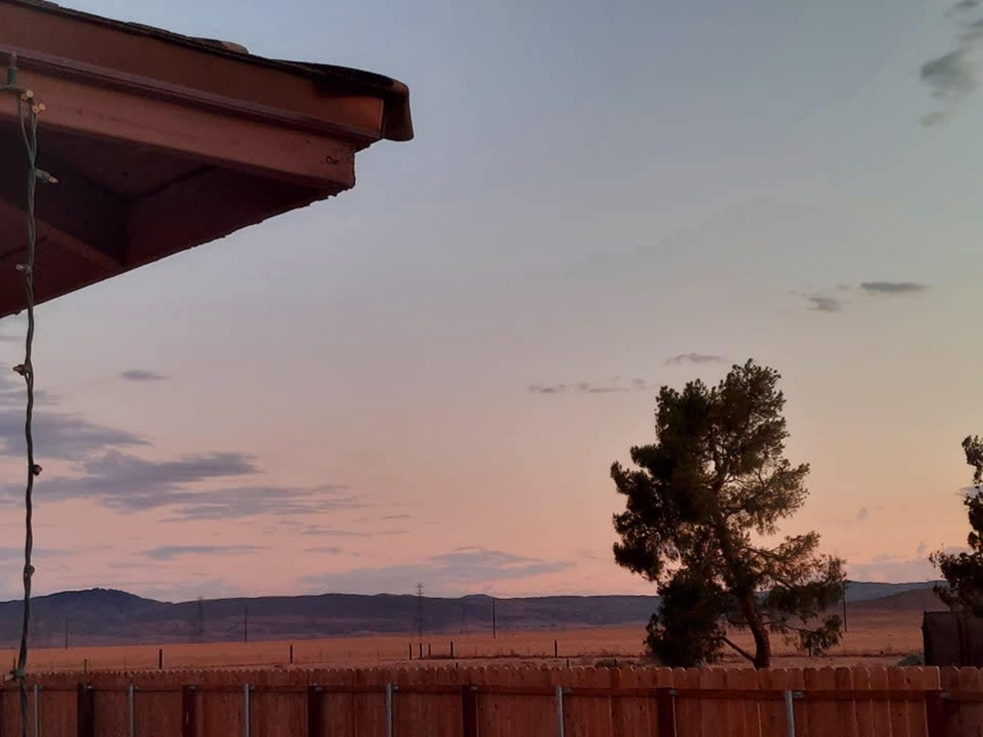 A wooden fence encloses a view of a desert landscape at dusk, featuring rolling hills and a single tree under a pastel sky.