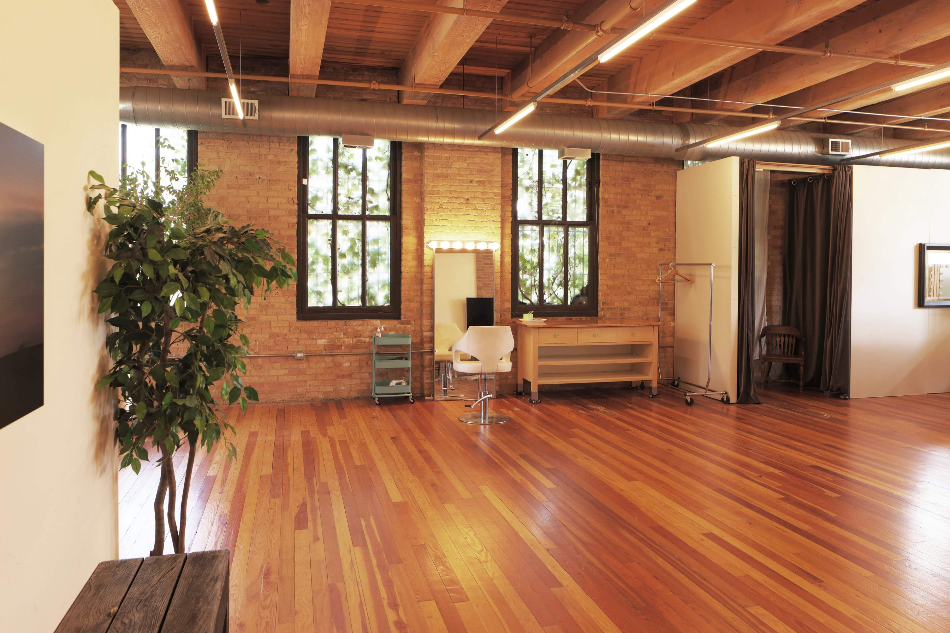 A spacious room with wooden floors, large windows, and minimal furnishings, including a chair, a dressing table, and a plant.