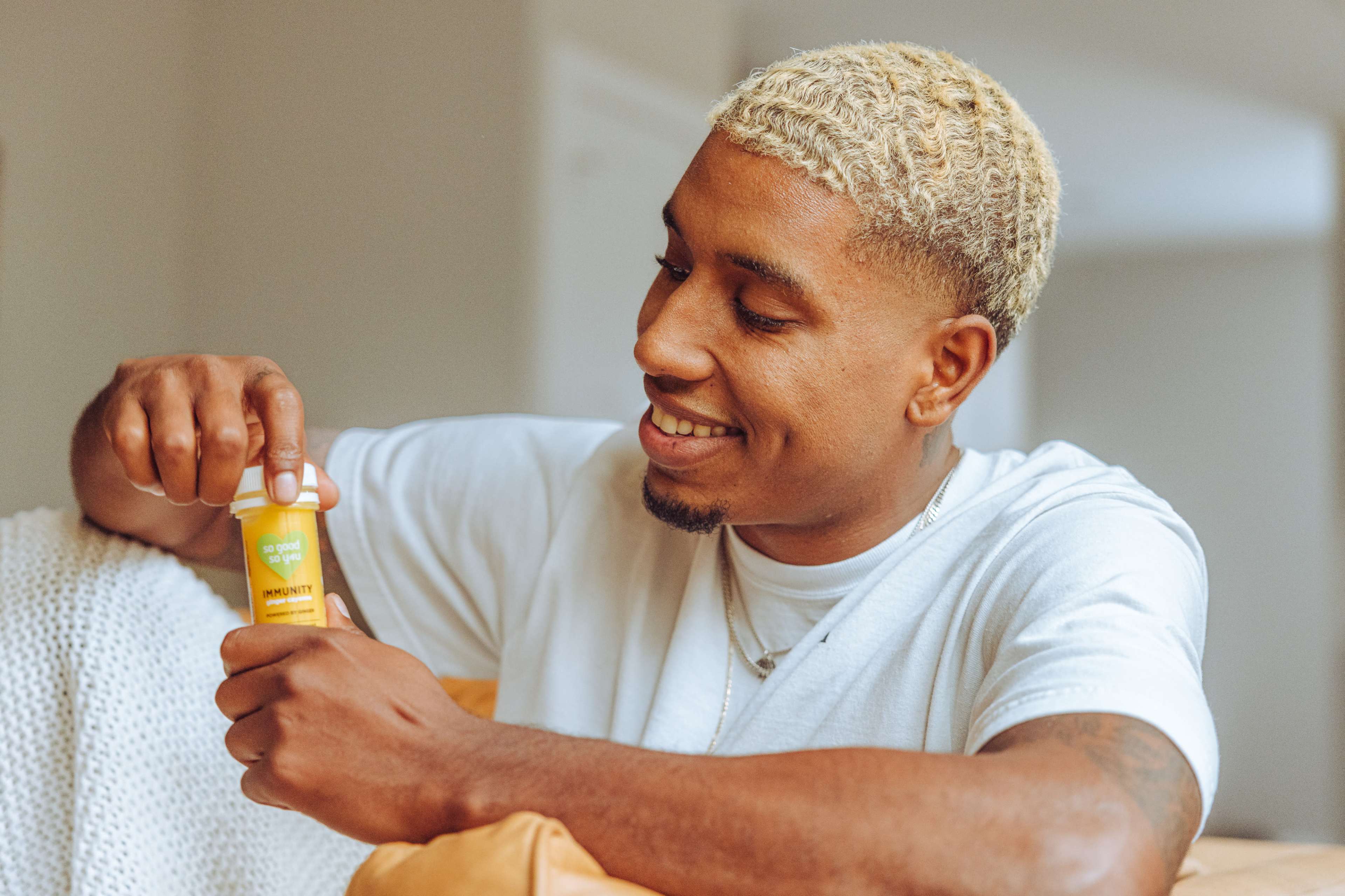 A man is sitting on a couch, smiling as he opens a yellow bottle of a product.