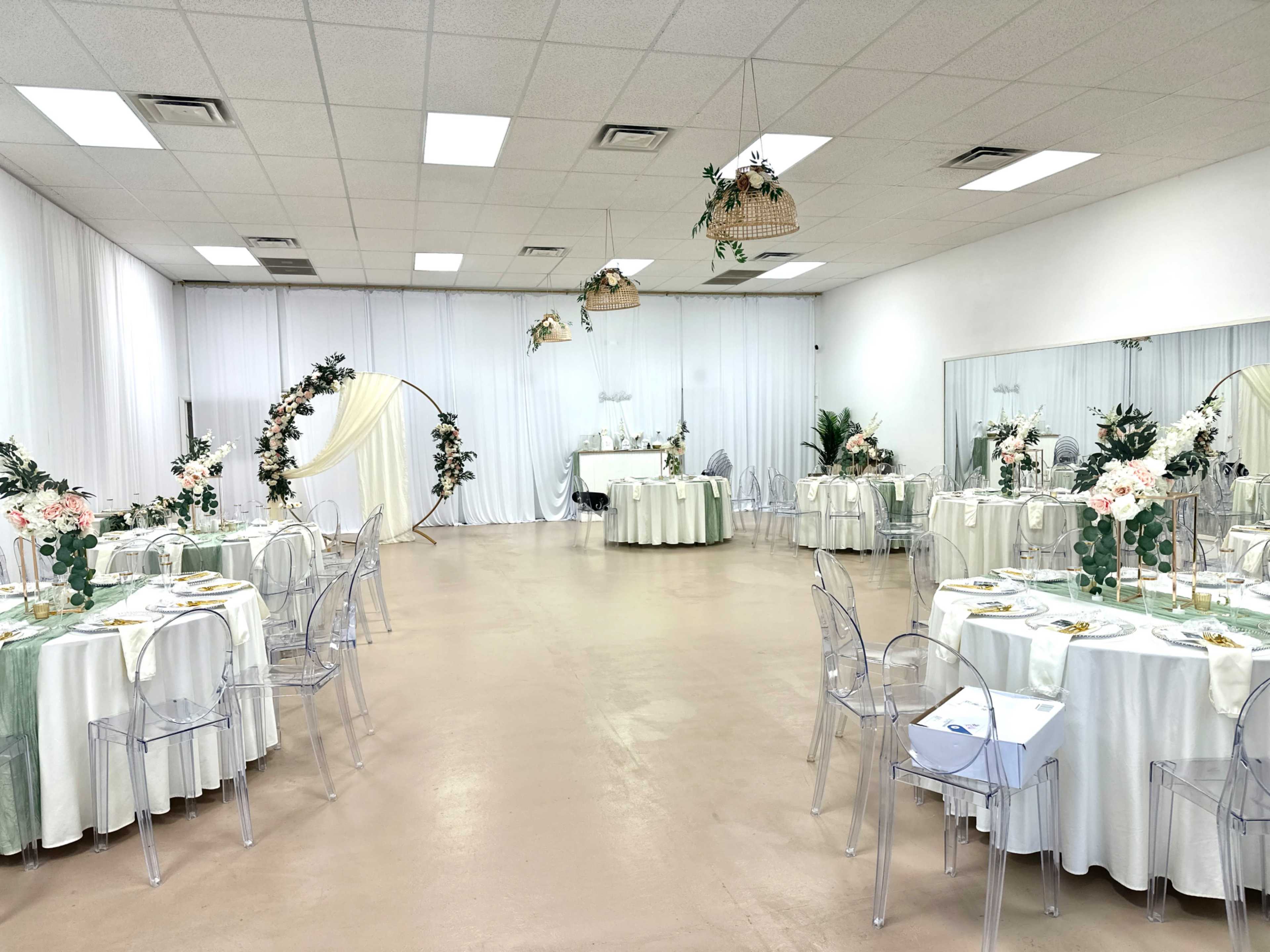 The image shows a spacious event venue arranged with several round tables covered in white and light green tablecloths, surrounded by clear chairs, and decorated with floral centerpieces.
