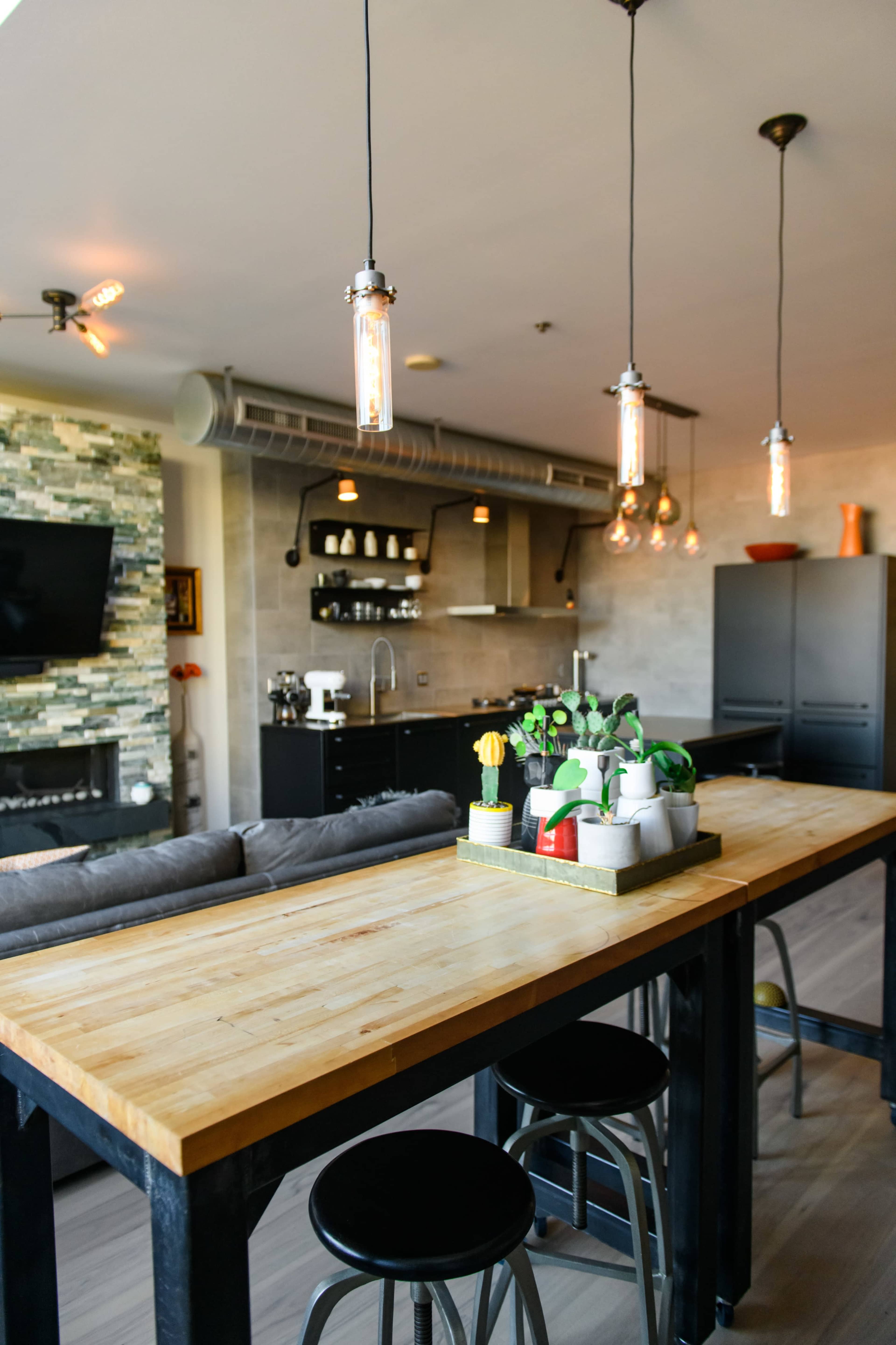 A modern kitchen features a wooden dining table with metal stools, pendant lighting, and a stone accent wall.