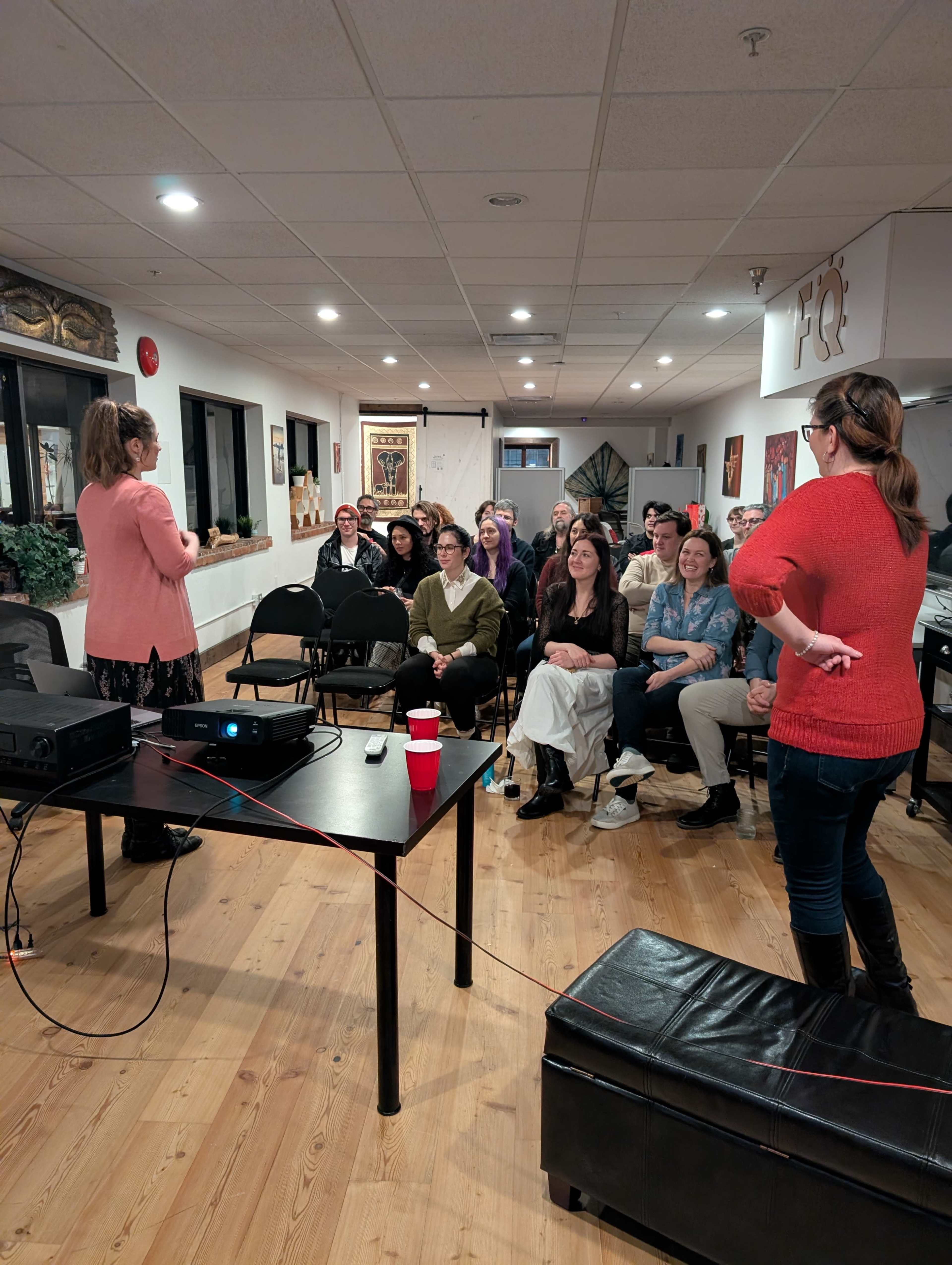A speaker stands in front of an audience seated in a small event space, with a projector and screen set up to the side.