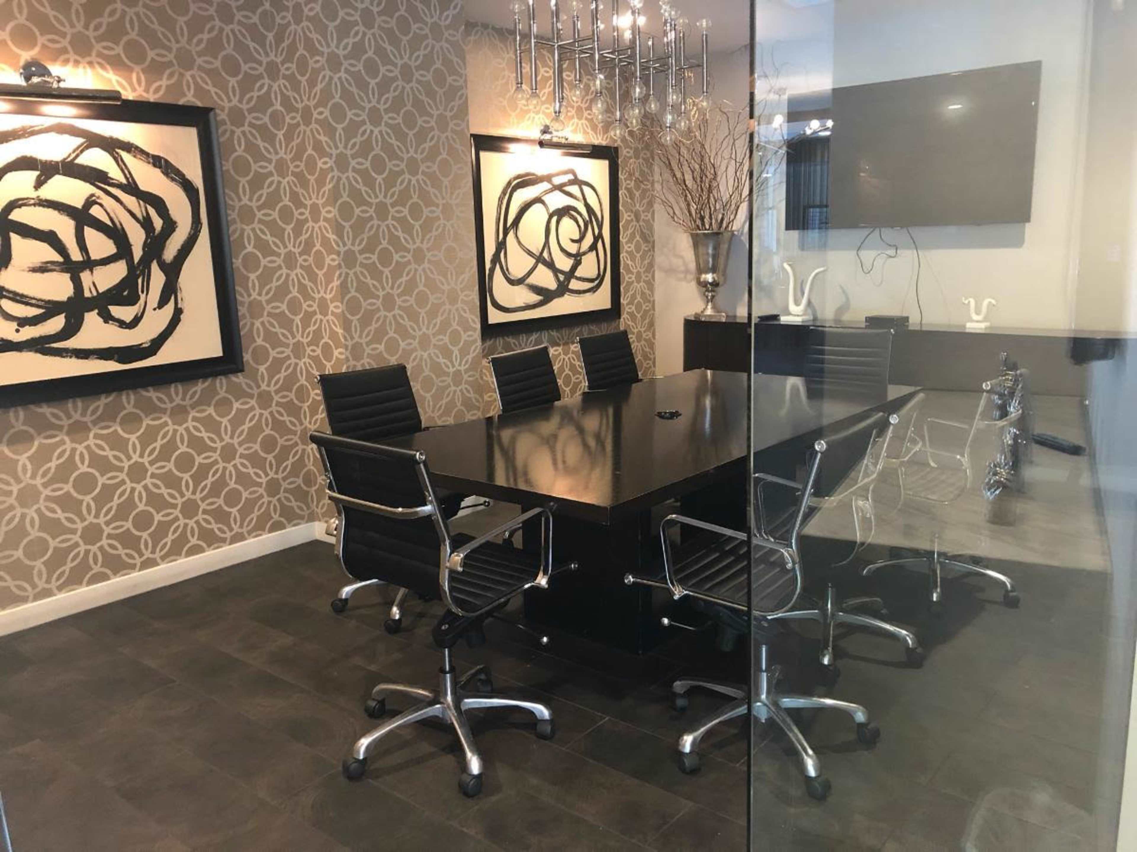 A modern conference room features a large black table surrounded by silver office chairs, with abstract wall art and a large screen on one side.