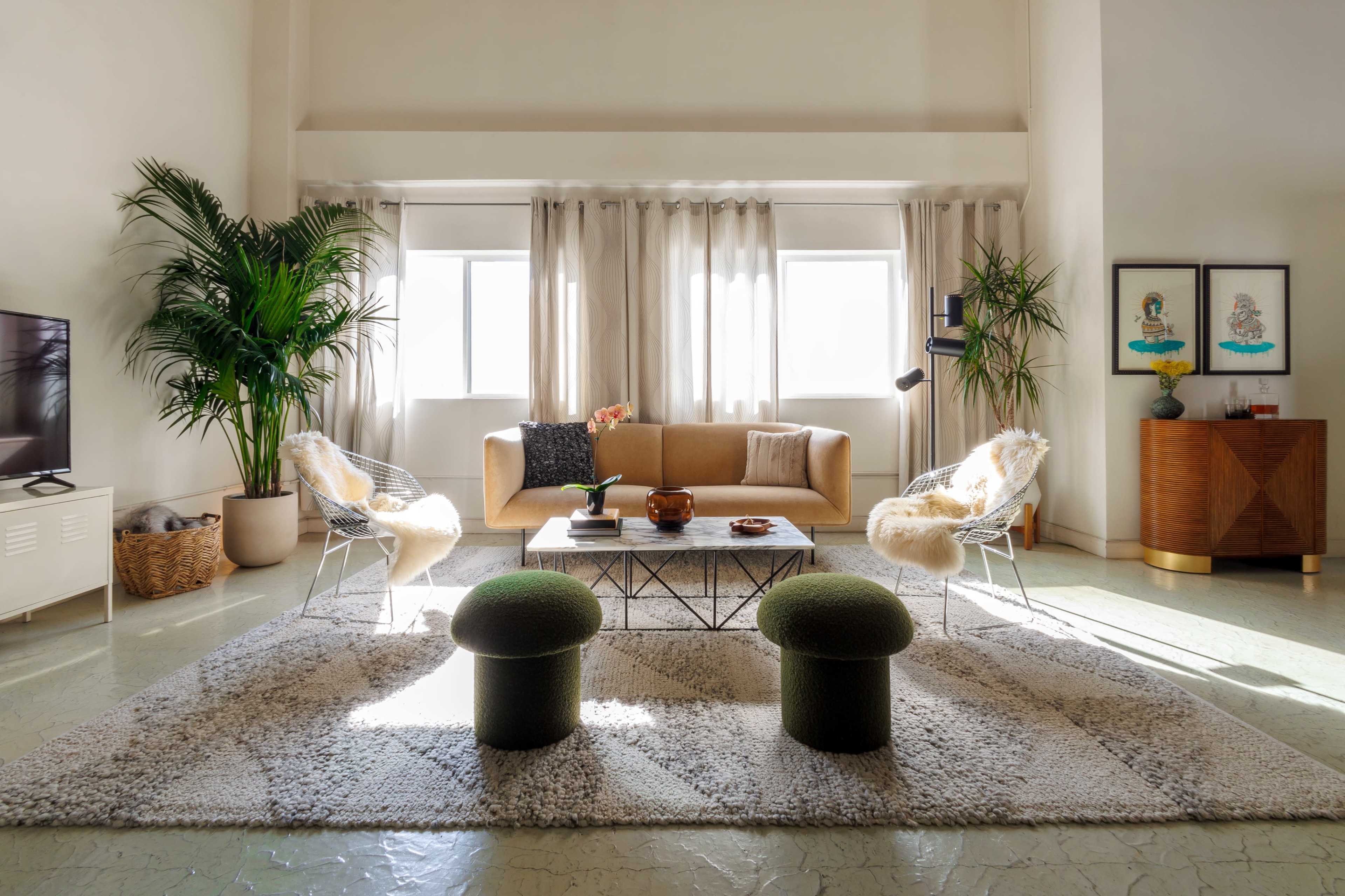 A bright living room features a tan sofa, two green stools, and a coffee table, surrounded by light-colored walls and large windows.